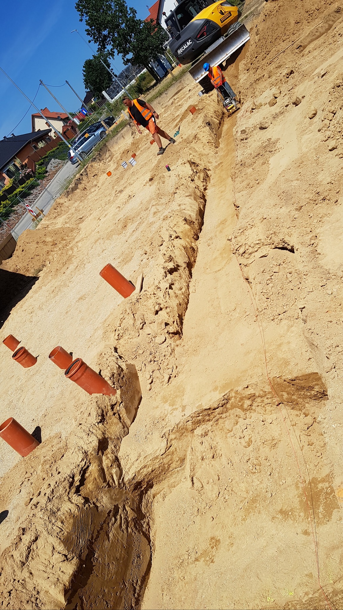 Wykop w ziemi z widocznymi rurami kanalizacyjnymi, pracownik ubija ziemię w wykopie, a w tle stoi minikoparka.