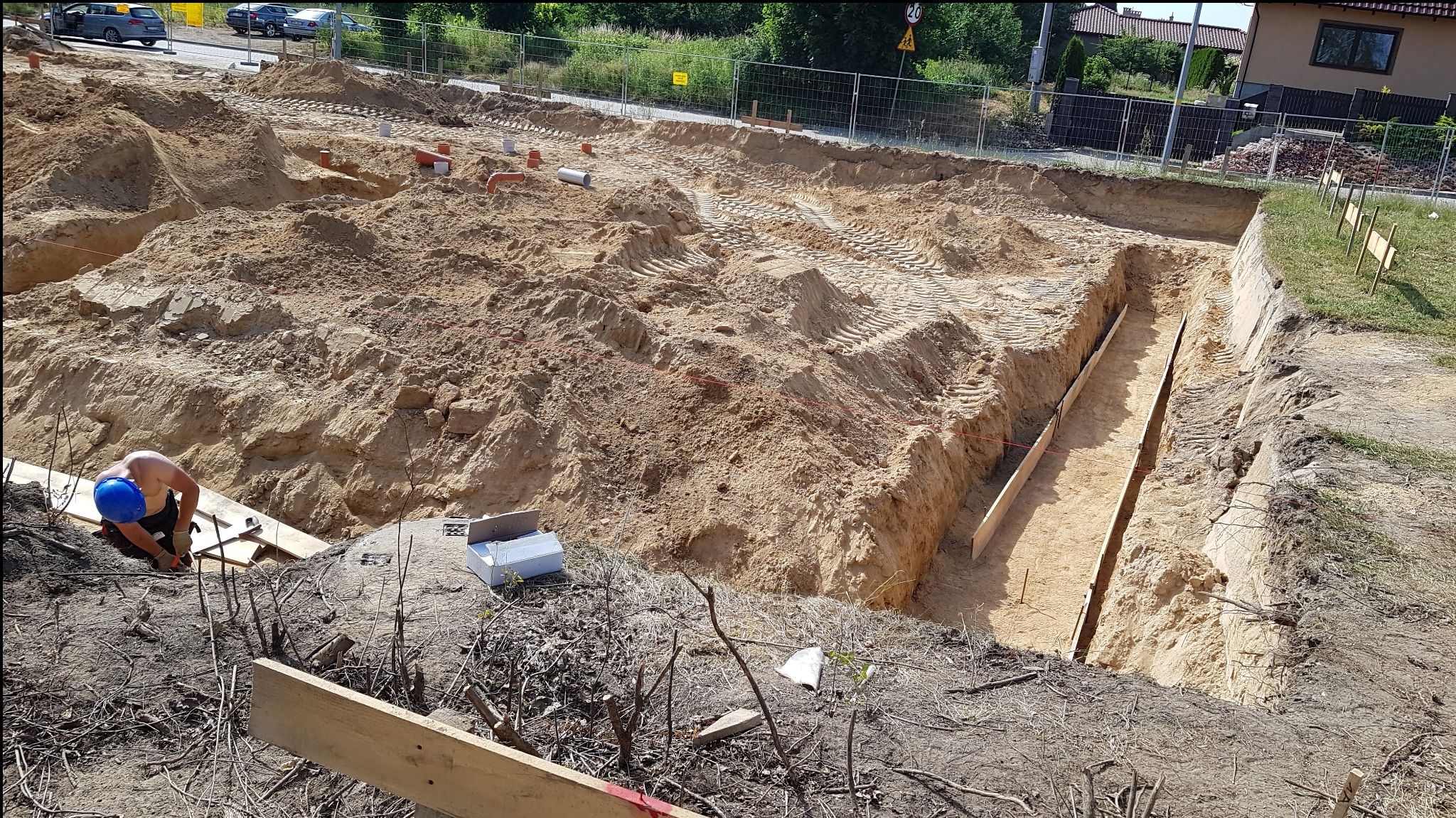 Przygotowanie wykopu pod fundamenty domu jednorodzinnego: widoczne szalunki w wykopie, pracownik w niebieskim kasku przy pracy, rury kanalizacyjne w piasku.