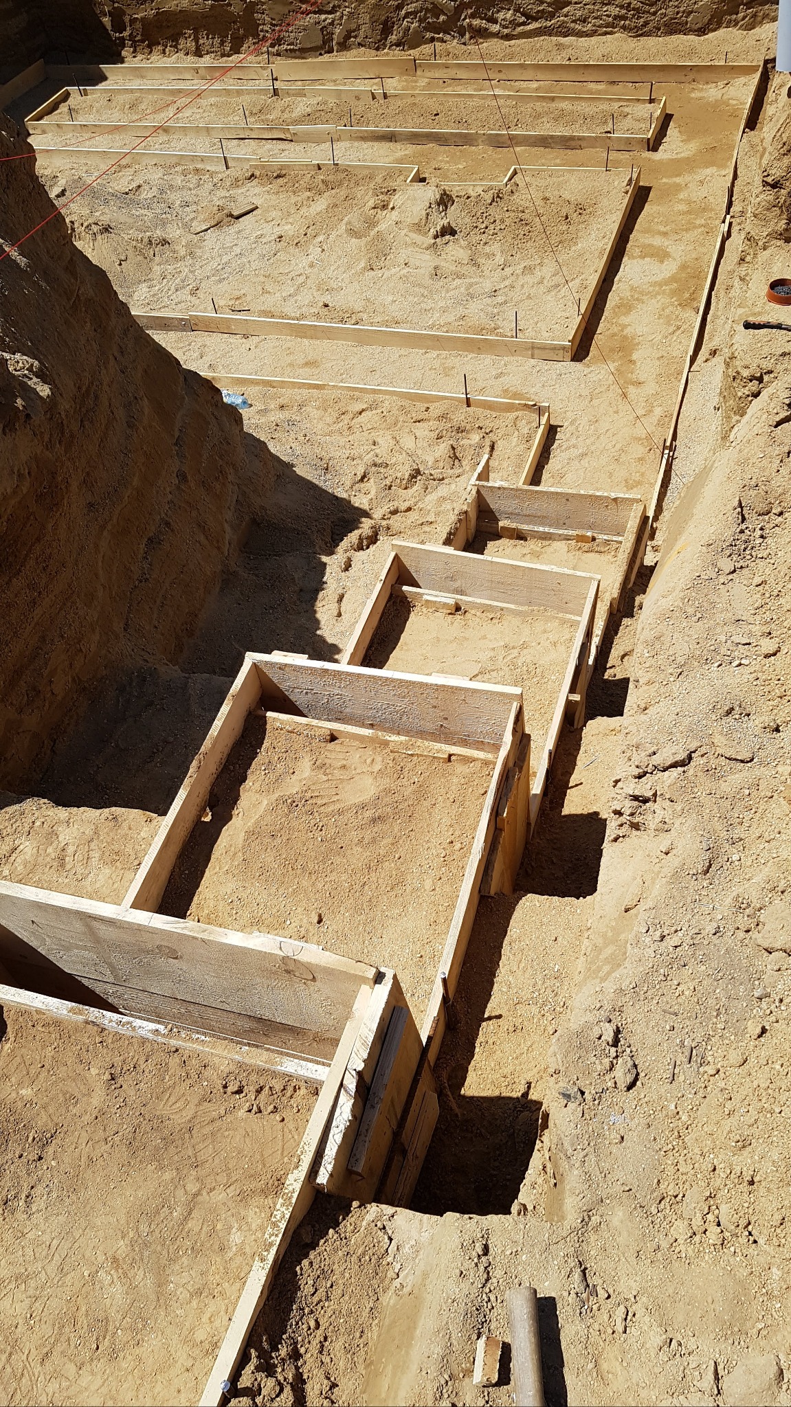 Szeroki kadr z góry na wykop pod fundamenty schodkowe z drewnianym szalunkiem wypełnionym piaskiem, widoczne sznurki wyznaczające obrys przyszłej konstrukcji.