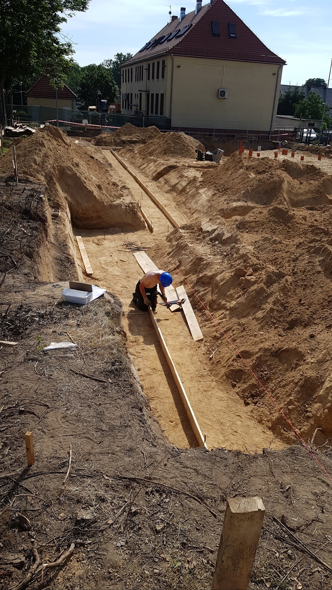 Pracownik w niebieskim kasku, bez koszulki, przykuca w wykopie fundamentowym, montując drewniane szalunki; w tle budynek mieszkalny i hałdy ziemi.