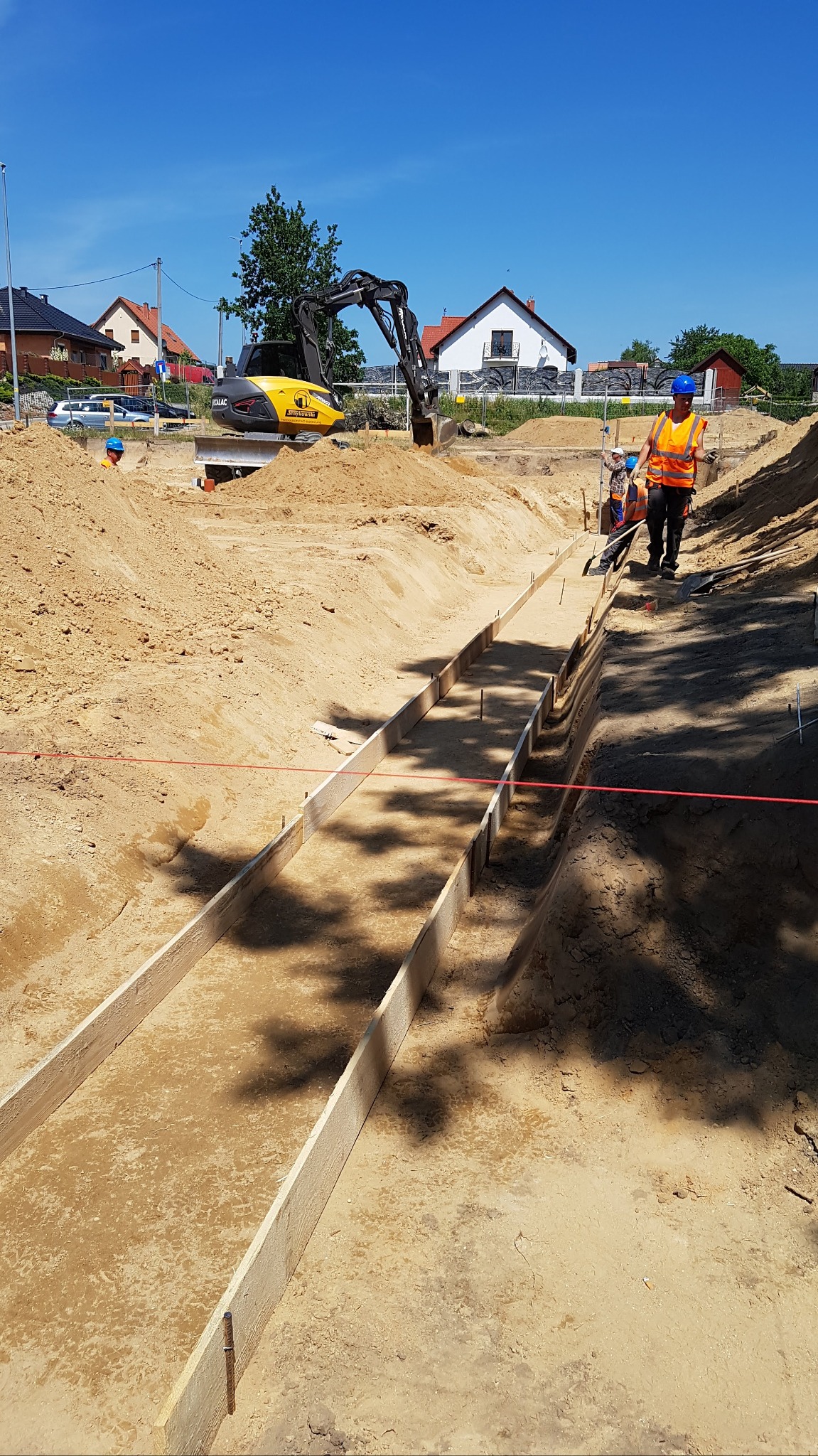 Przygotowanie wykopu pod fundamenty: żółta koparka w tle, szalunki z desek w wykopie, pracownicy w odblaskowych kamizelkach i kaskach.