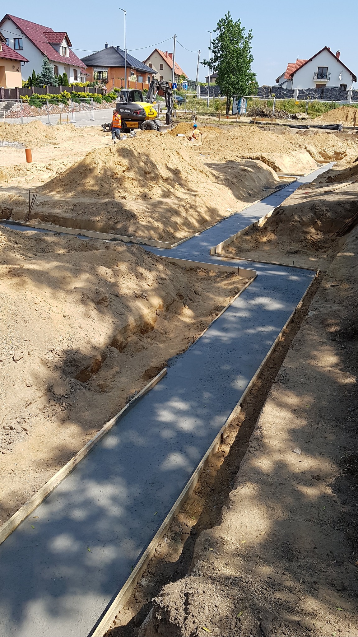 Wylane betonowe ławy fundamentowe w wykopach, z widocznym szalunkiem drewnianym, na tle placu budowy z koparką i domami jednorodzinnymi w tle.