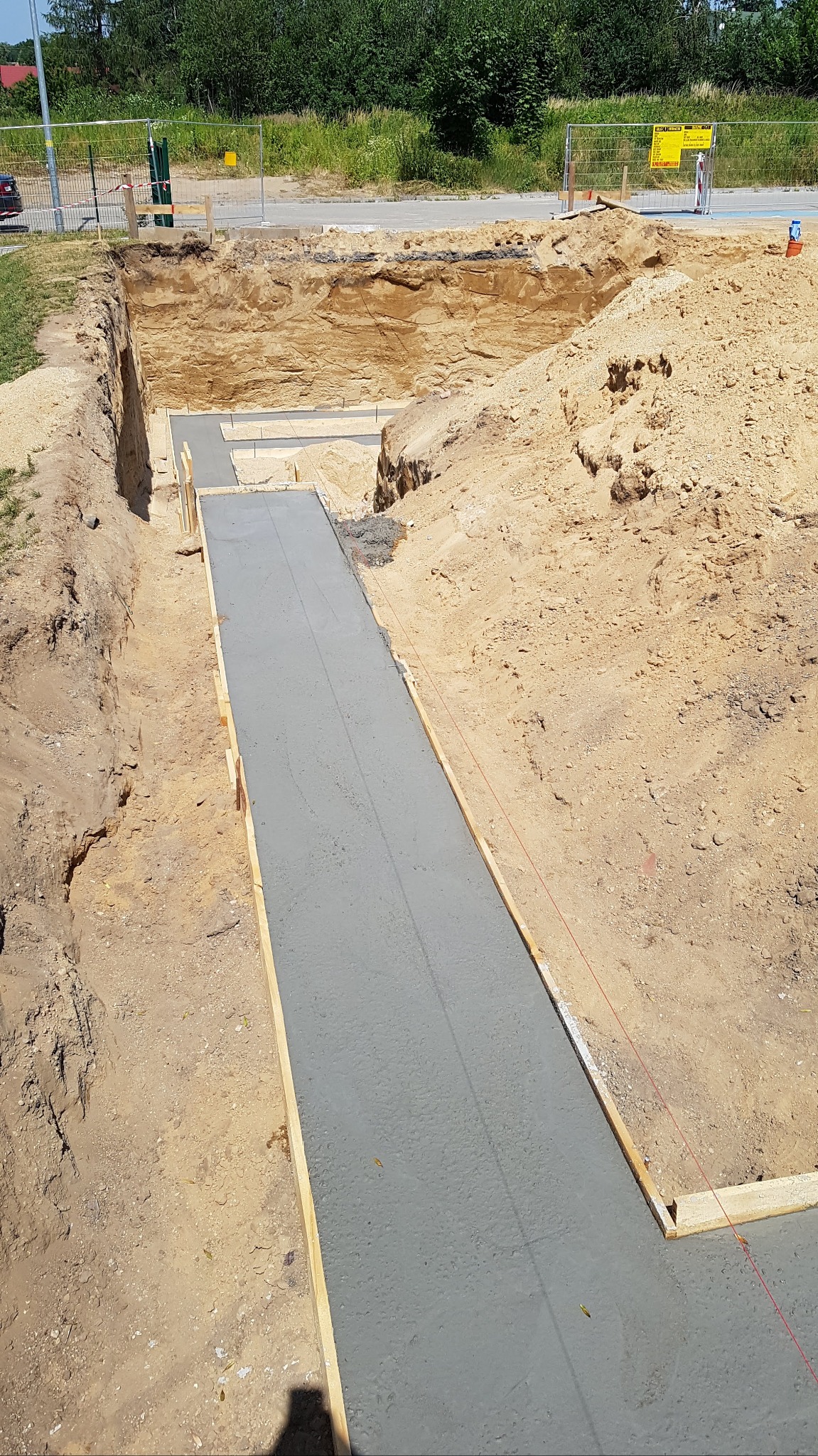 Wylany betonowy fundament w wykopie z drewnianym szalunkiem, widoczne naprężone sznurki wyznaczające linie, ziemia wokół wykopu.