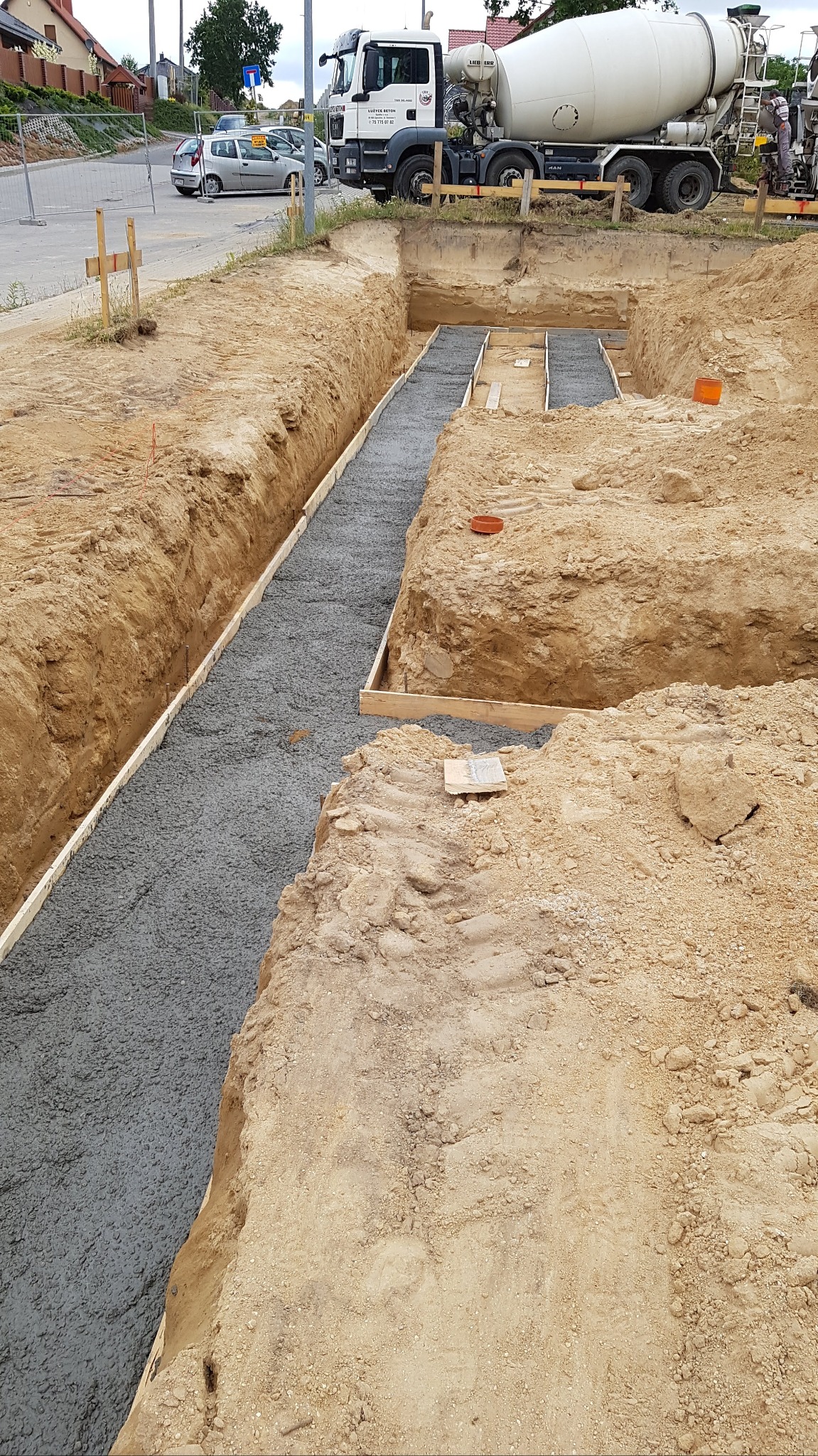 Świeżo wylany beton w wykopie pod fundamenty, z widocznym szalunkiem i betoniarką w tle, na tle ulicy z zaparkowanymi samochodami.
