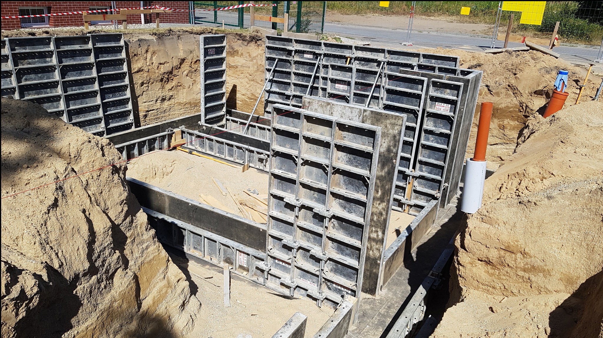 Szczegółowy widok wykopu pod fundamenty z użyciem szalunków systemowych, widoczny piasek na dnie wykopu i rury kanalizacyjne wystające z ziemi.