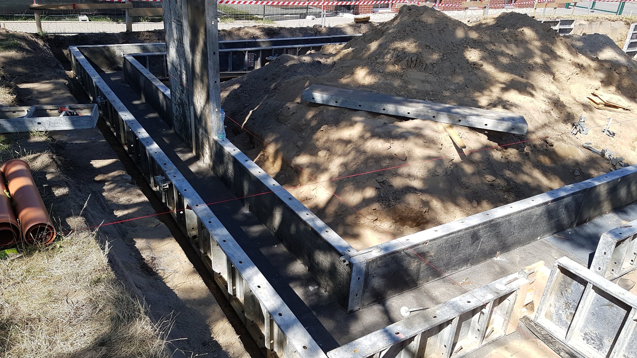 Szare szalunki fundamentowe, z widoczną stertą piasku i wykopem, zabezpieczone czerwoną linką, z rurami PCV w tle, na budowie w słoneczny dzień.