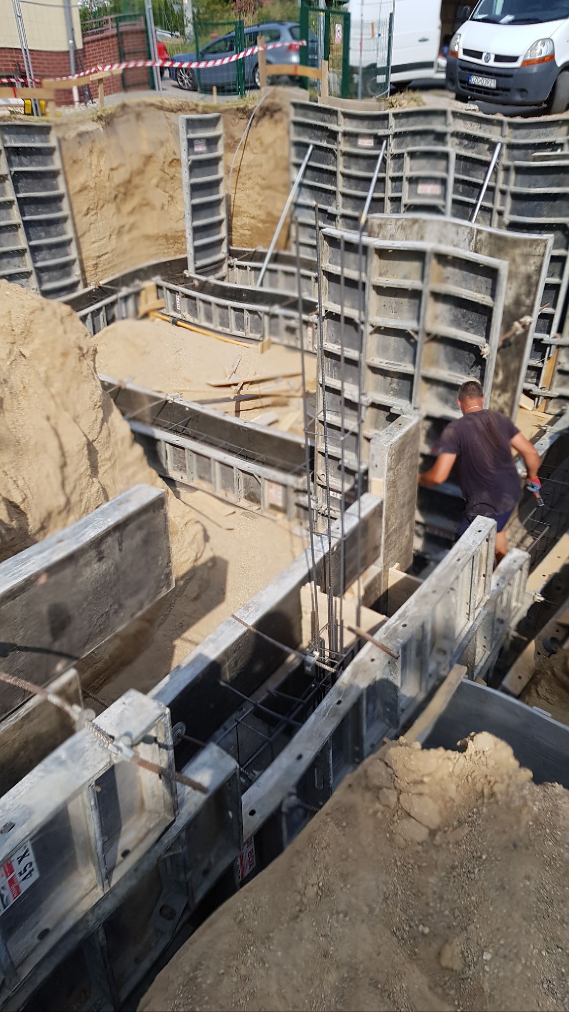 Wykop fundamentów z szalunkami systemowymi i zbrojeniem, widoczny pracownik w trakcie pracy, na drugim planie zaparkowane samochody.