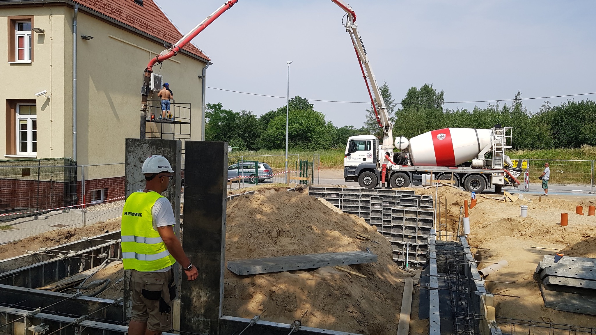 Budowa fundamentów: widok na szalunek, zbrojenie i kopiec piasku, w tle betonomieszarka z wysięgnikiem i pracownicy nadzorujący wylewanie betonu.