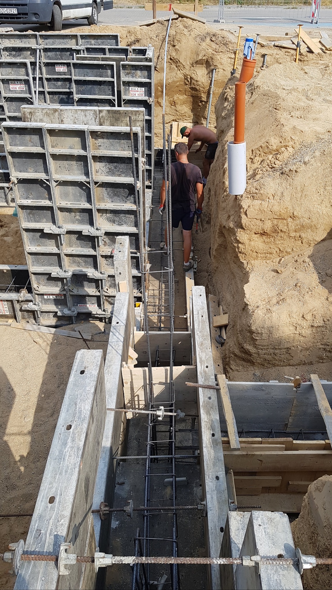 Przygotowanie zbrojenia i szalunków pod fundament, widoczni dwaj pracownicy w wykopie, jeden z nich w zielonej czapce, w tle widoczne rury instalacyjne i samochód dostawczy.