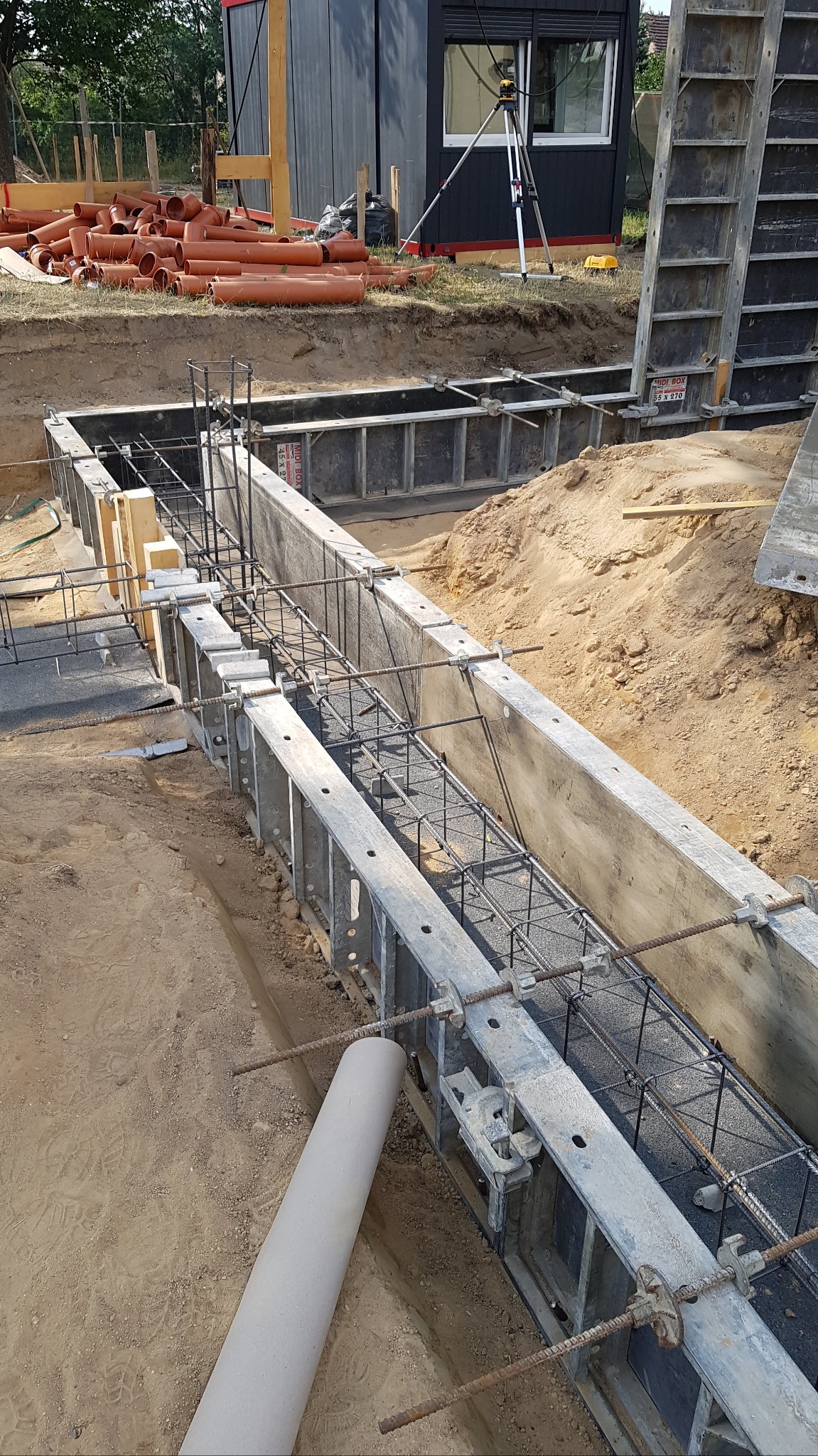 Metalowe szalunki zbrojone stalą, przygotowane do zalania betonem, widok z góry na placu budowy z rurami PCV w tle i budką biurową.