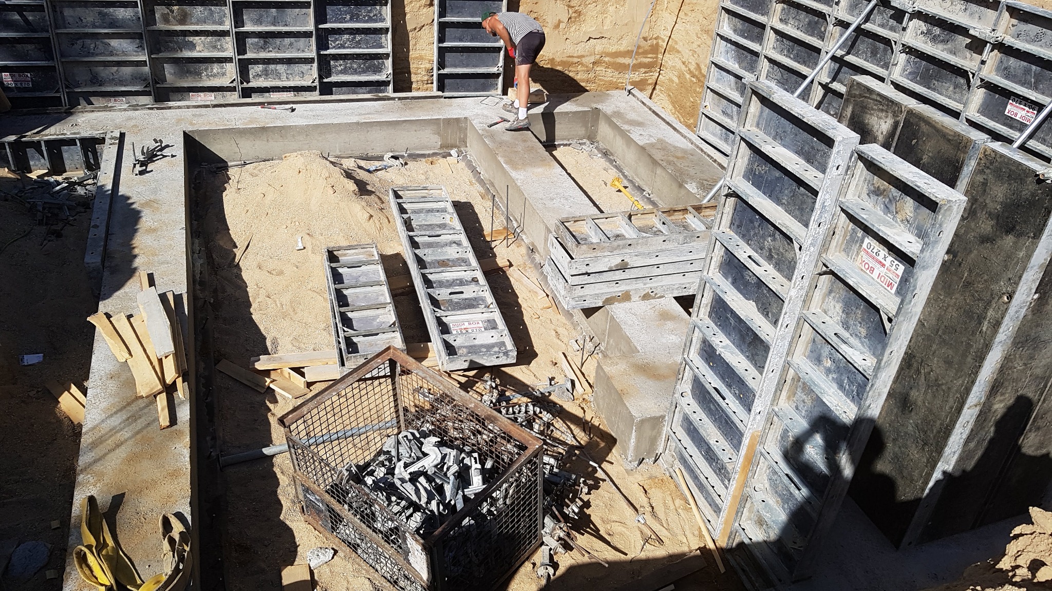 Budowa fundamentów: widok z góry na wykop z betonową wylewką, szalunki systemowe, narzędzia, oraz pracownika w koszulce w paski i czarnych spodenkach.