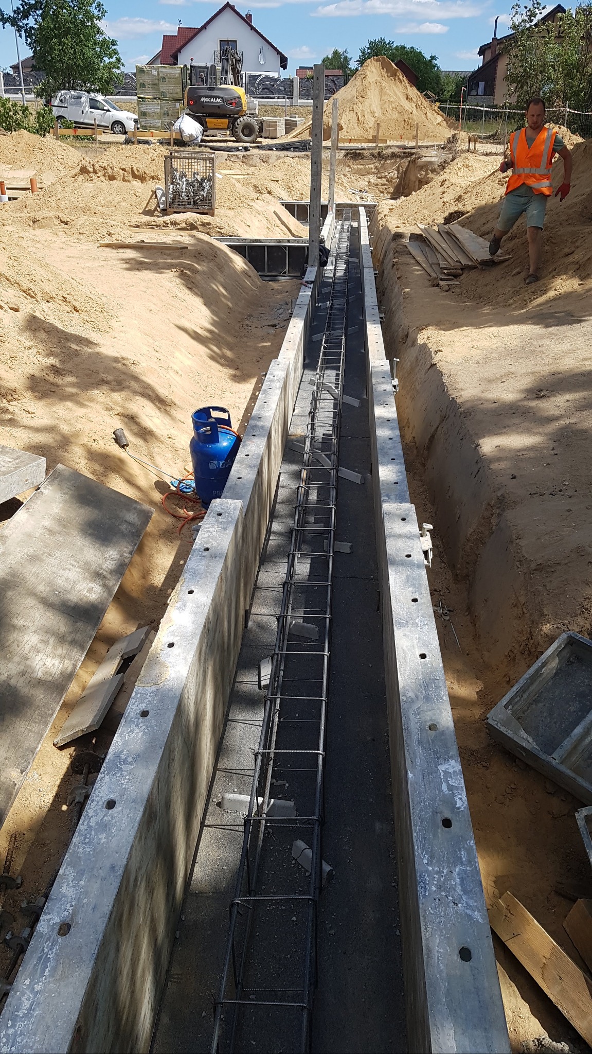 Wykop pod fundamenty z ułożonym zbrojeniem w szalunku, na budowie widoczna butla gazowa, koparka i pracownik w kamizelce odblaskowej.