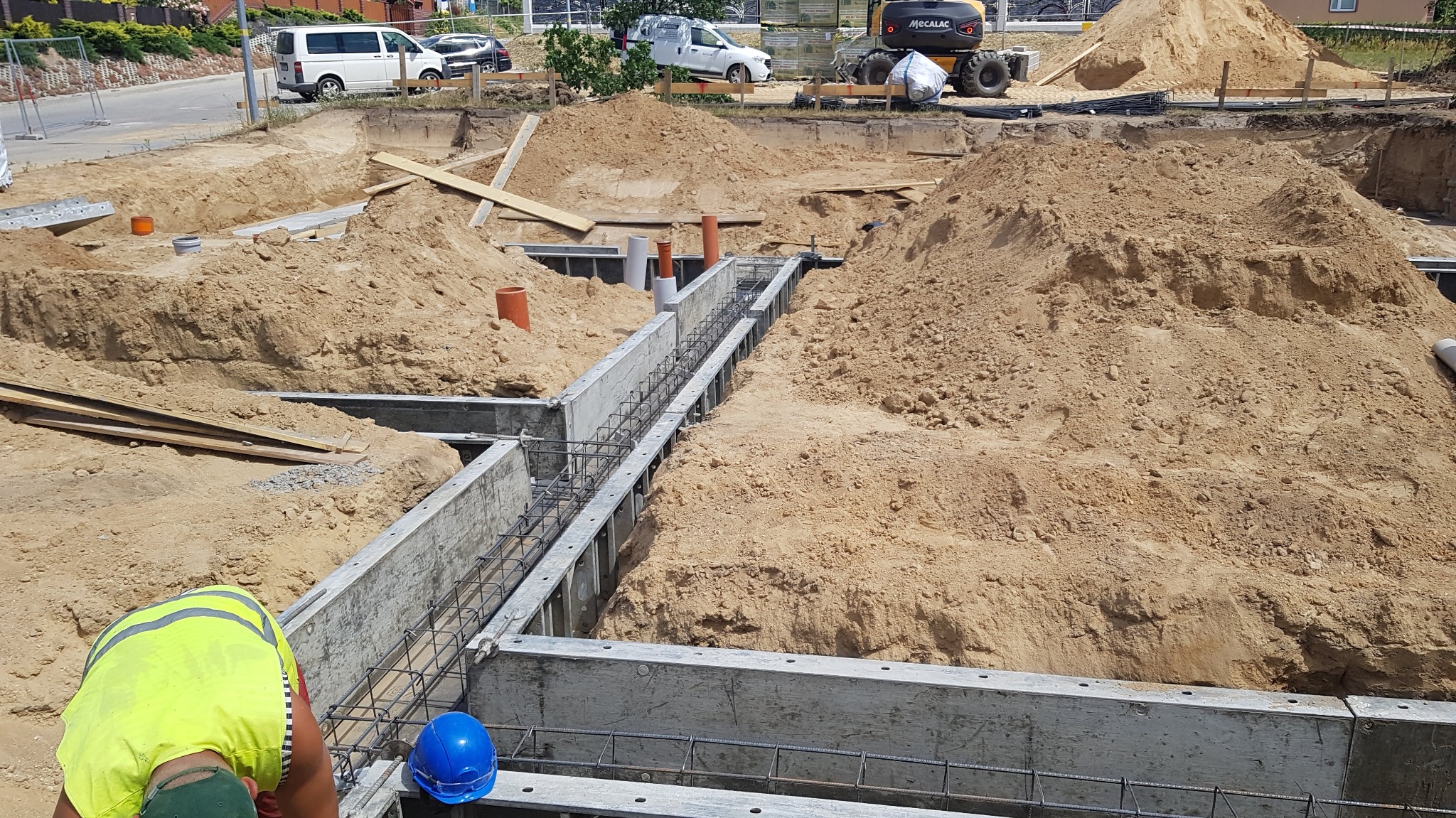 Wykop pod fundamenty z szalunkiem, zbrojeniem i widoczną stertą piasku, pracownik w żółtej kamizelce oraz niebieski kask na pierwszym planie, w tle zaparkowane samochody.
