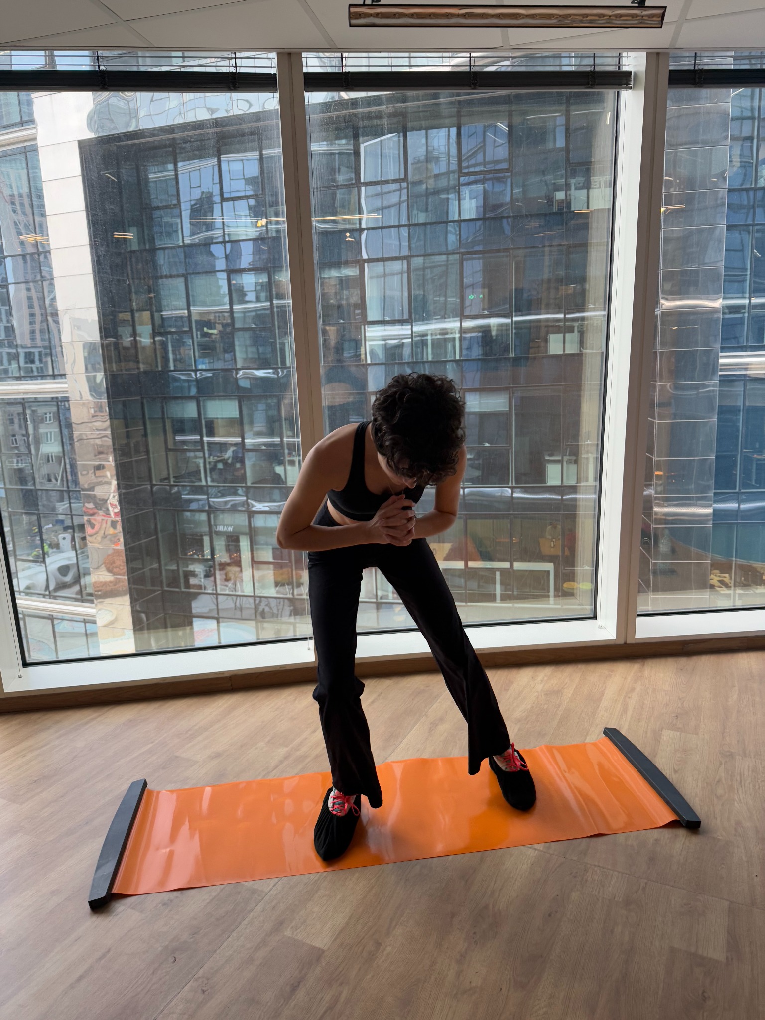 Kobieta w sportowym stroju ćwiczy na pomarańczowej macie do ślizgania, z widokiem na nowoczesne budynki przez okno.