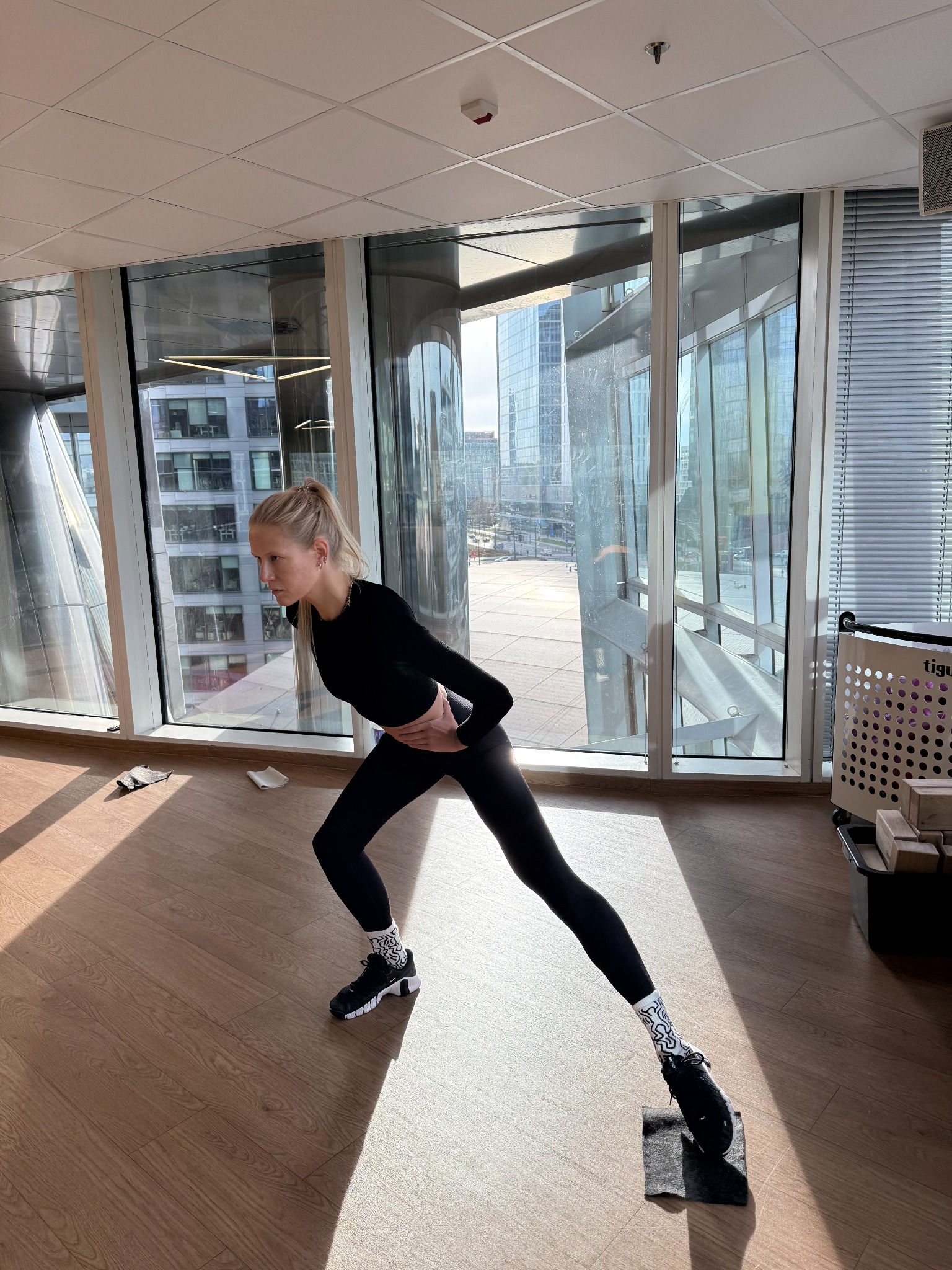 Kobieta w czarnym stroju sportowym wykonuje wypad w bok na małej platformie w przestronnym pomieszczeniu z widokiem na miasto.