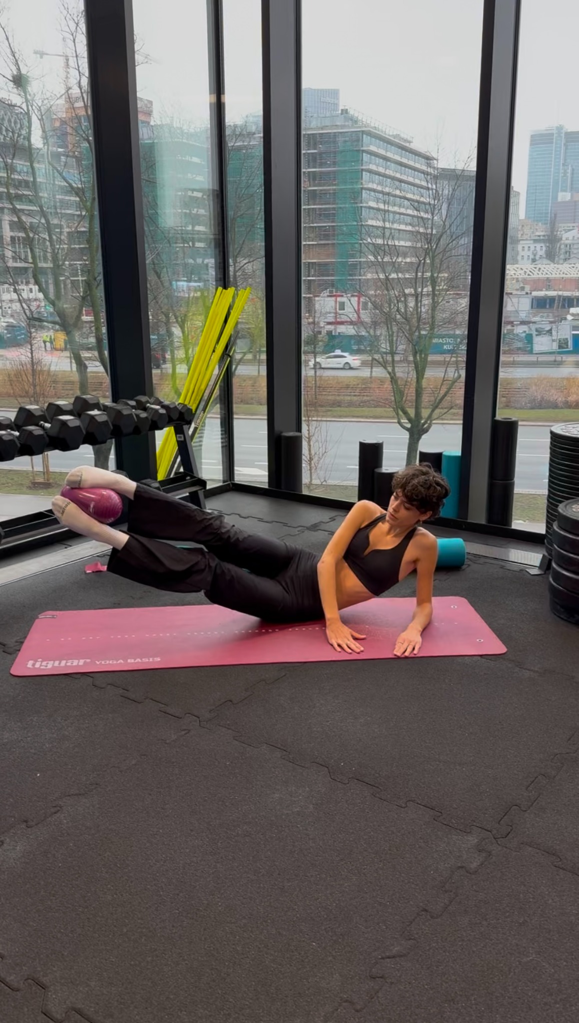 Kobieta w czarnym stroju sportowym ćwiczy unoszenie nóg z piłką między stopami, leżąc na różowej macie do jogi w siłowni z widokiem na miasto.