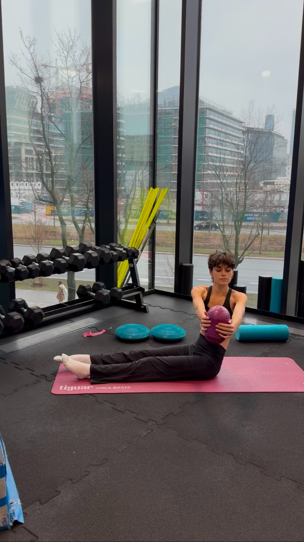 Kobieta w czarnym stroju sportowym wykonuje ćwiczenie z fioletową piłką na różowej macie w przestronnym, dobrze oświetlonym studio fitness z widokiem na miasto.