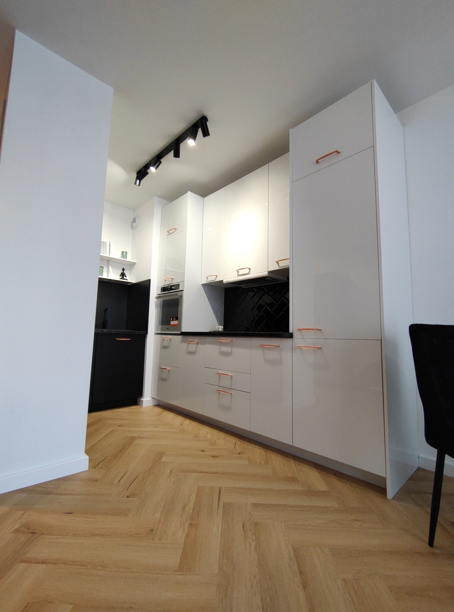 Nowoczesna kuchnia z białymi, błyszczącymi frontami, miedzianymi uchwytami i czarną ścianą nad blatem, oświetlona reflektorami, z parkietem ułożonym w jodełkę.