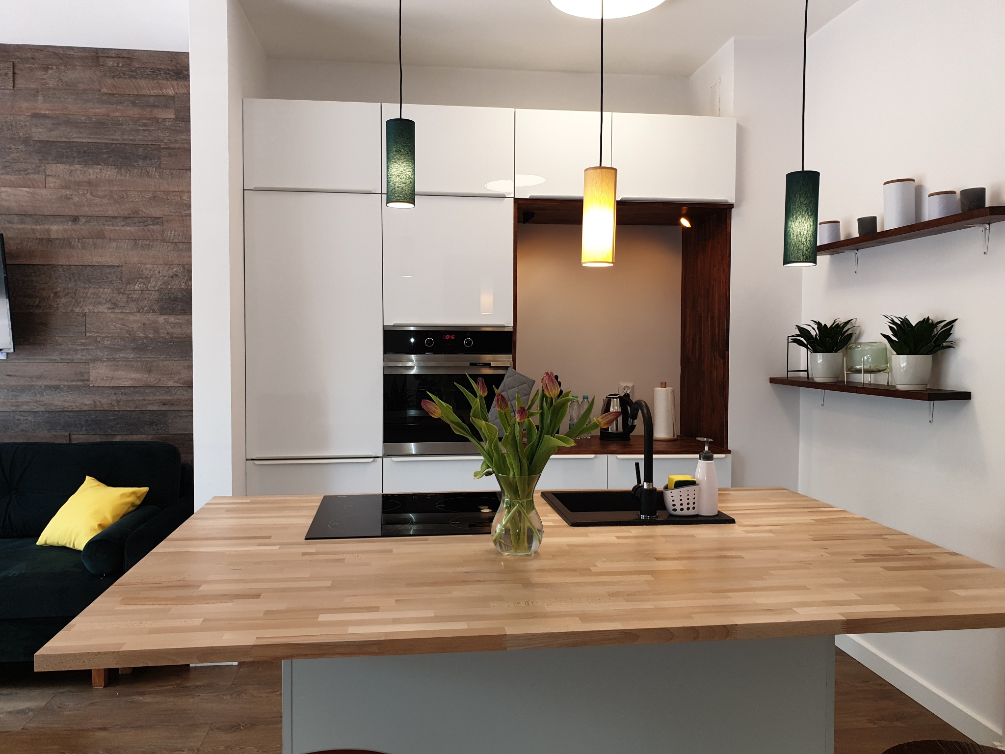 Nowoczesna kuchnia z białymi frontami, drewnianym blatem wyspy i wiszącymi lampami o cylindrycznych abażurach w odcieniach zieleni i żółci, uzupełniona o drewnianą ścianę w tle i zieloną sofę...