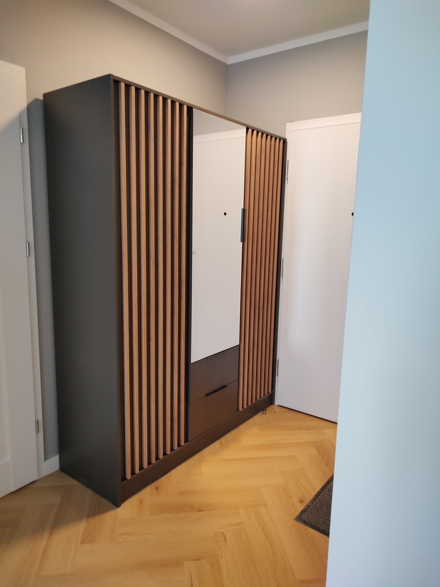 Nowoczesna, dwudrzwiowa szafa z lustrem i drewnianymi lamelami na frontach, umieszczona w przedpokoju z parkietem w jodełkę.
