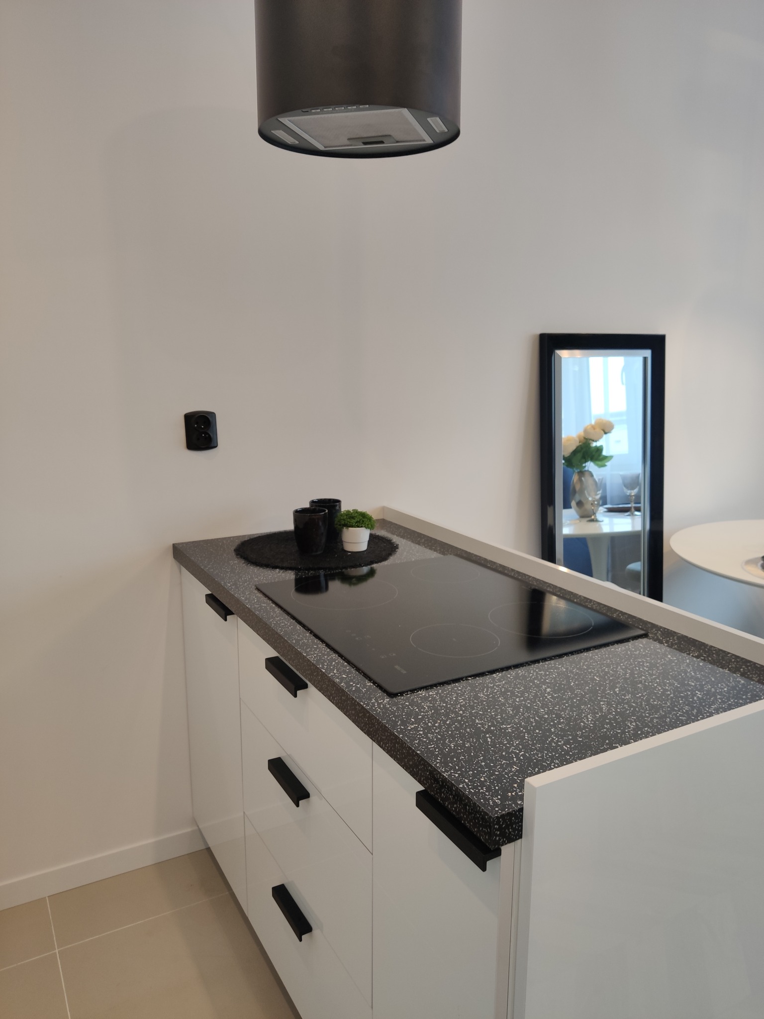 Fragment nowoczesnej kuchni z białymi szafkami, czarnym blatem lastryko, płytą indukcyjną i okrągłym okapem; dekoracje: czarne kubki i roślina w doniczce, odbicie lustrzane z kwiatami.