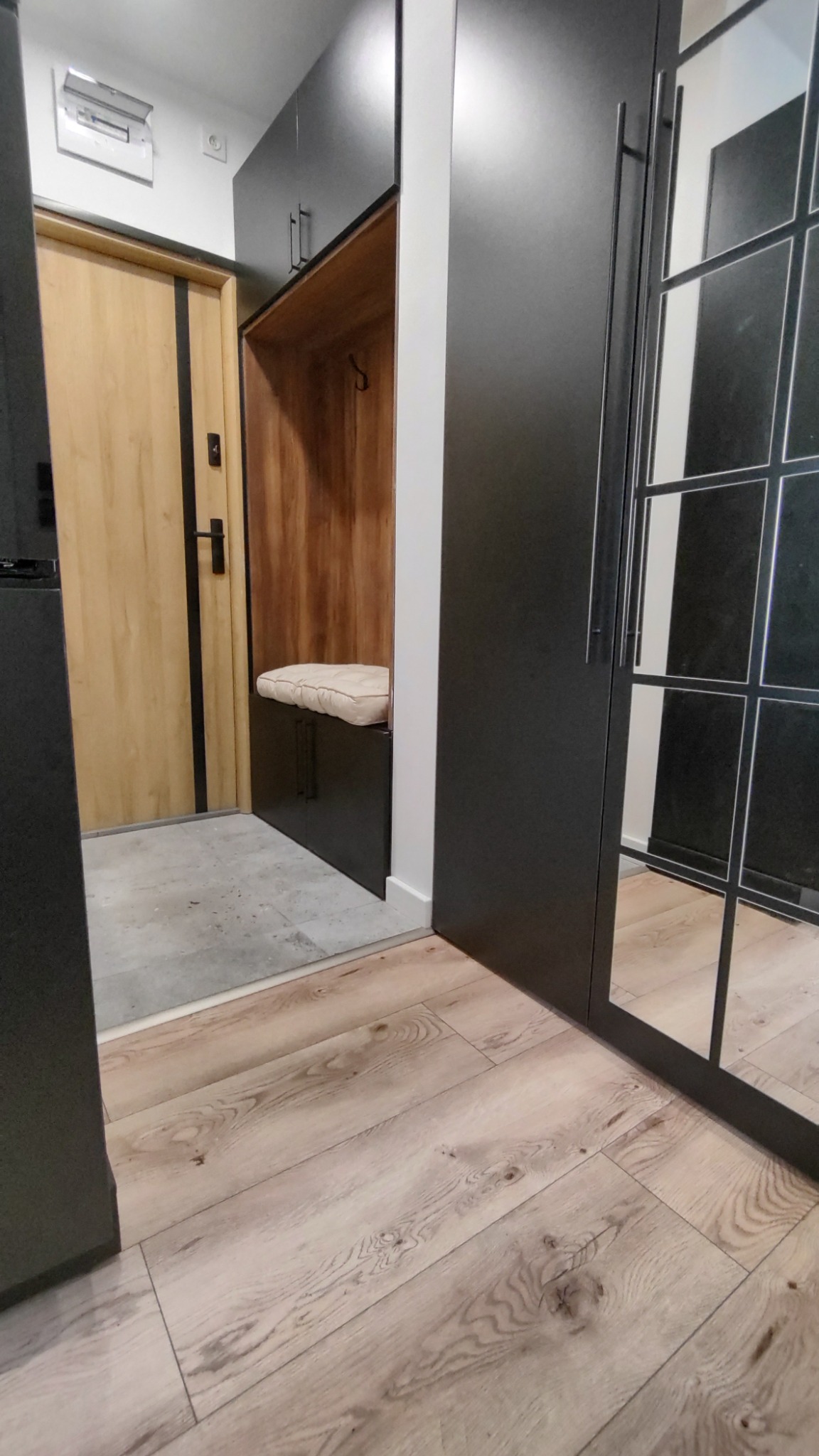 Przedpokój z zabudową meblową: szafa z lustrzanymi drzwiami w czarnej ramie, wnęka z siedziskiem i wieszakami, drewniana podłoga, widok z perspektywy wejścia.