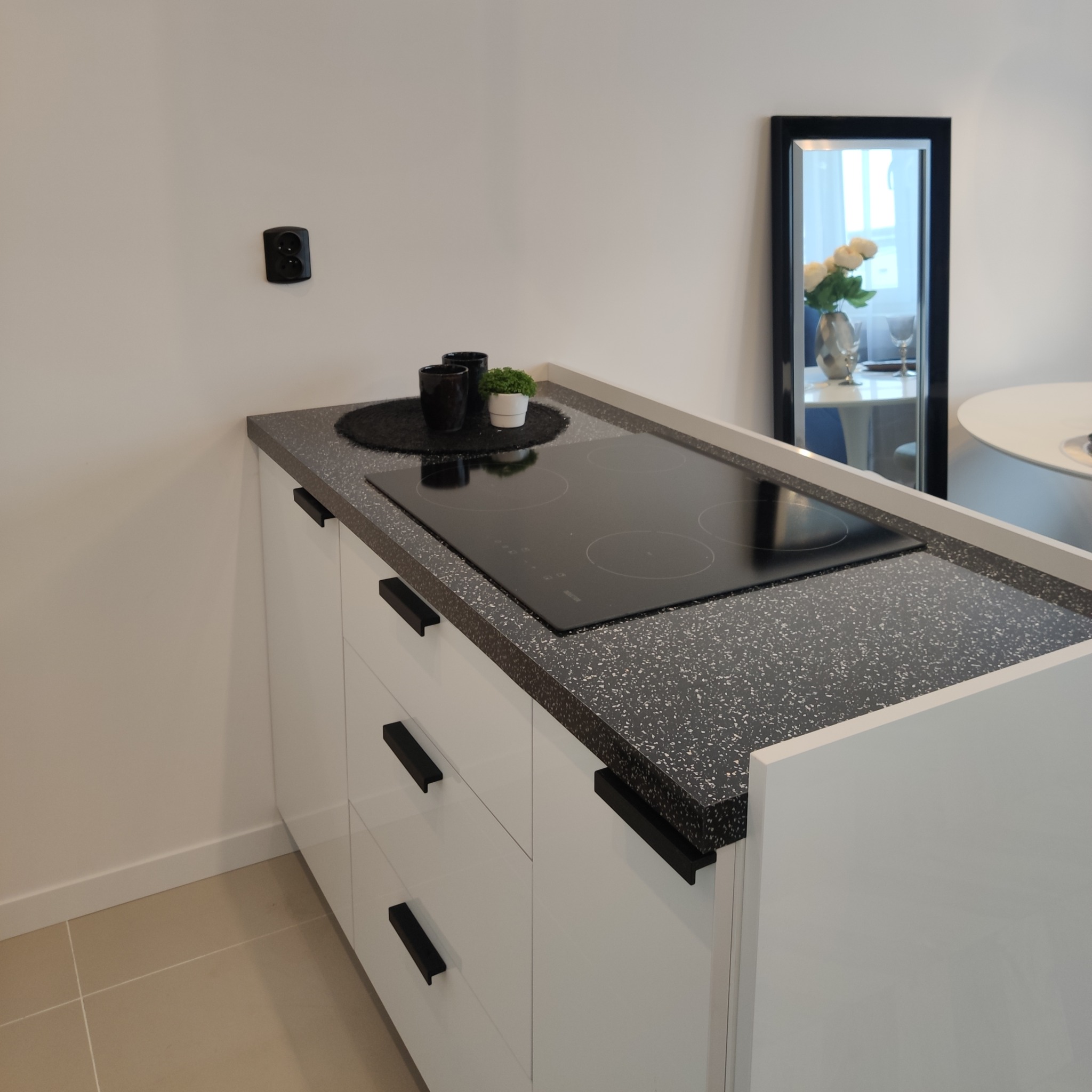 Minimalistyczna kuchnia: białe szafki z czarnymi uchwytami, blat z czarnego lastryko, płyta indukcyjna, czarne kubki i mała roślina doniczkowa na blacie, w tle lustro z odbiciem kwiatów w wazonie.