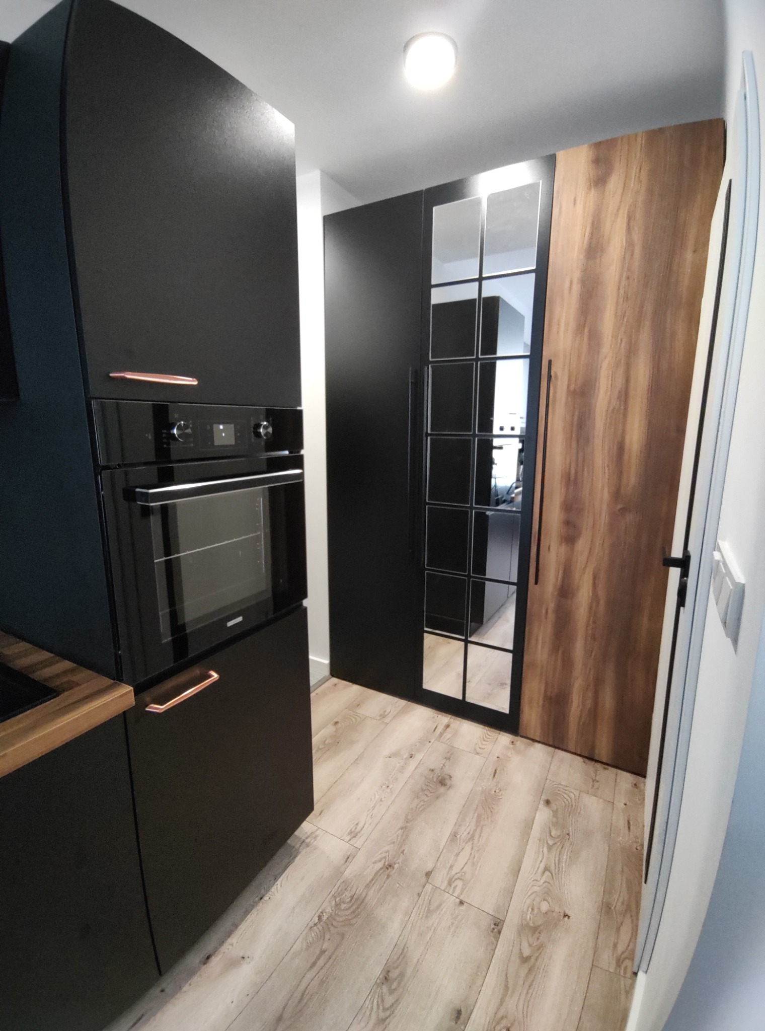 Nowoczesna kuchnia: czarne szafki z miedzianymi uchwytami, piekarnik w zabudowie, podłoga z paneli imitujących drewno, wysoka szafa z drewnianym frontem i czarnymi przeszkleniami.