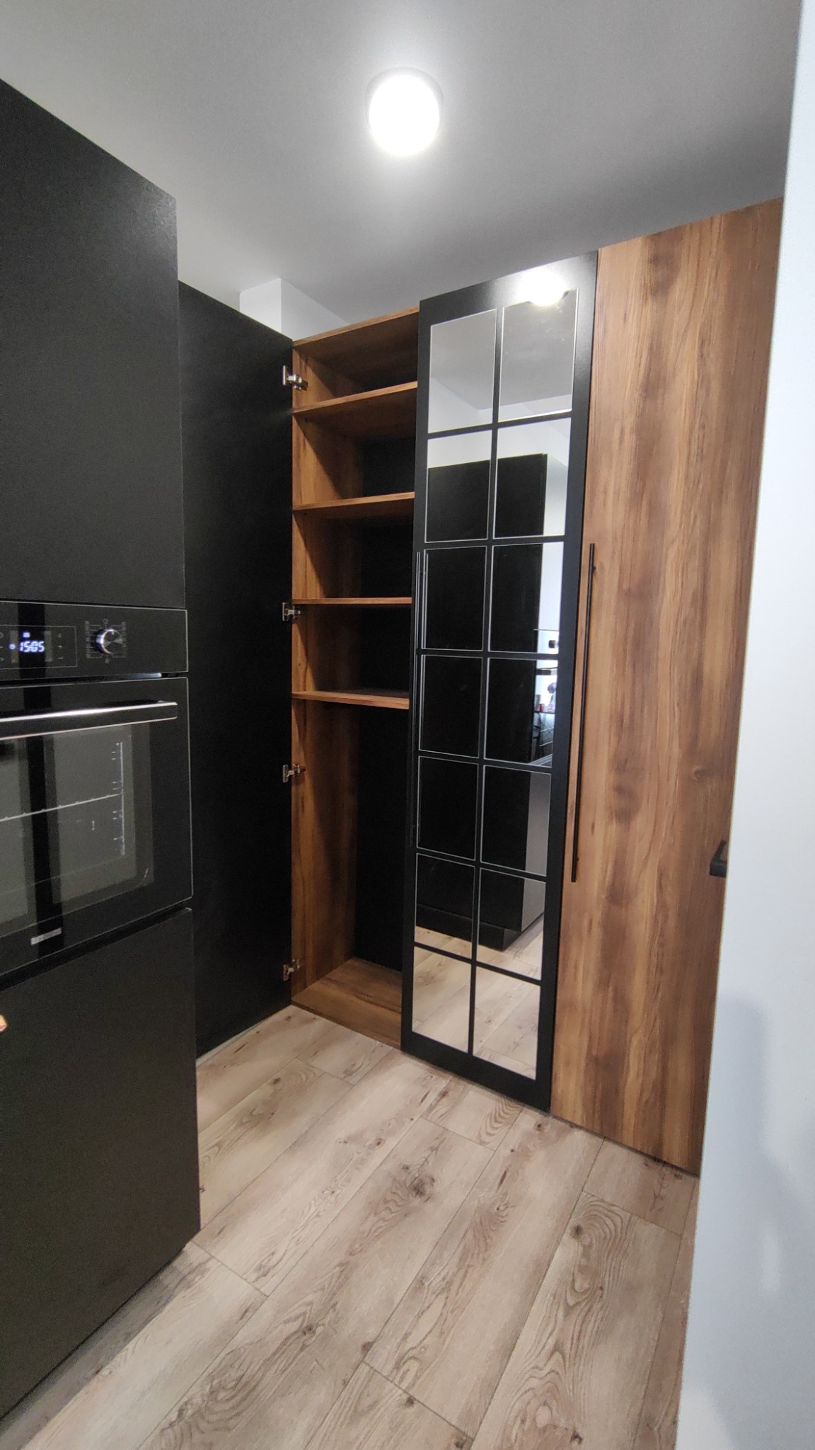 Częściowo otwarta, wysoka szafa z drewnianymi półkami i czarną, szklaną witryną, sąsiadująca z czarną zabudową kuchenną z piekarnikiem, na podłodze jasne panele.