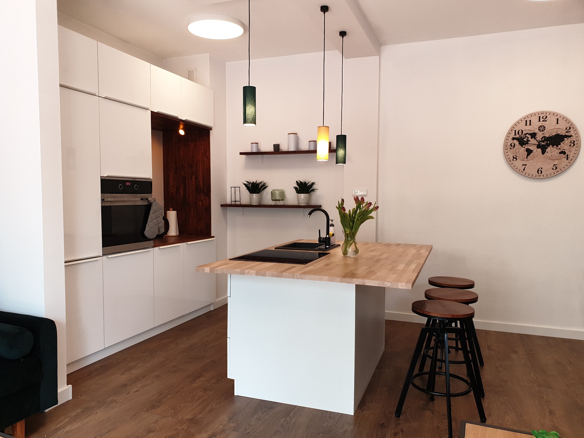 Nowoczesna kuchnia z białymi szafkami, drewnianym blatem wyspy i trzema hokerami, oświetlona wiszącymi lampami i z zegarem z mapą świata na ścianie.