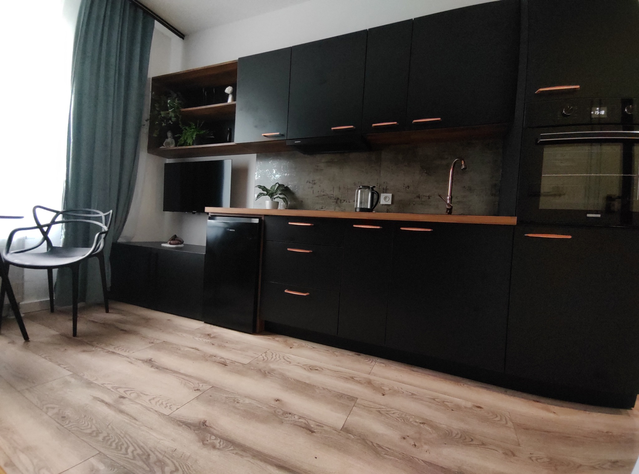 Nowoczesna kuchnia z czarnymi frontami, drewnianymi blatami i miedzianymi uchwytami, oświetlona naturalnym światłem z okna z turkusowymi zasłonami.