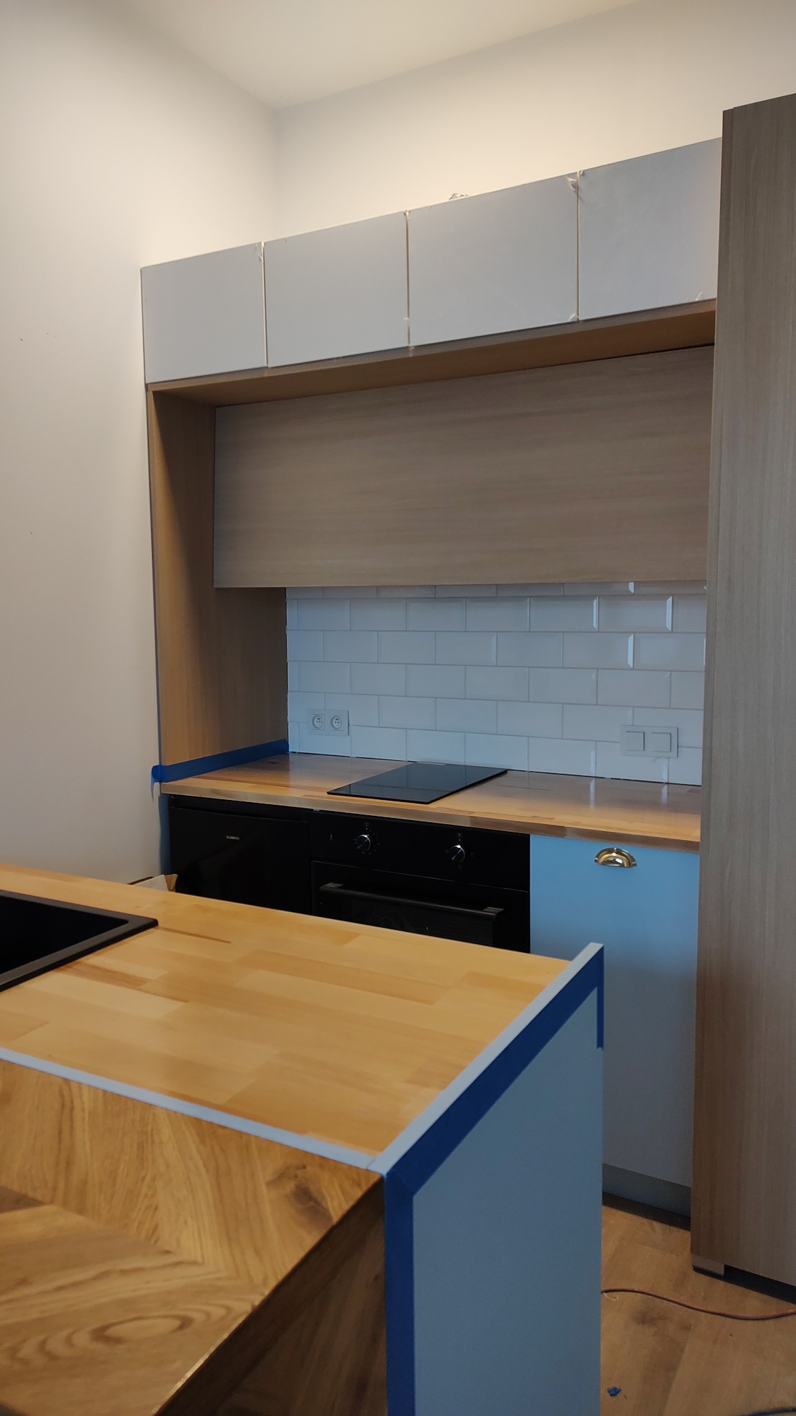 Fragment kuchni w trakcie montażu w Łodzi, z widoczną wyspą z blatem w jodełkę, płytą indukcyjną i białymi płytkami na ścianie, zabezpieczoną niebieską taśmą malarską.