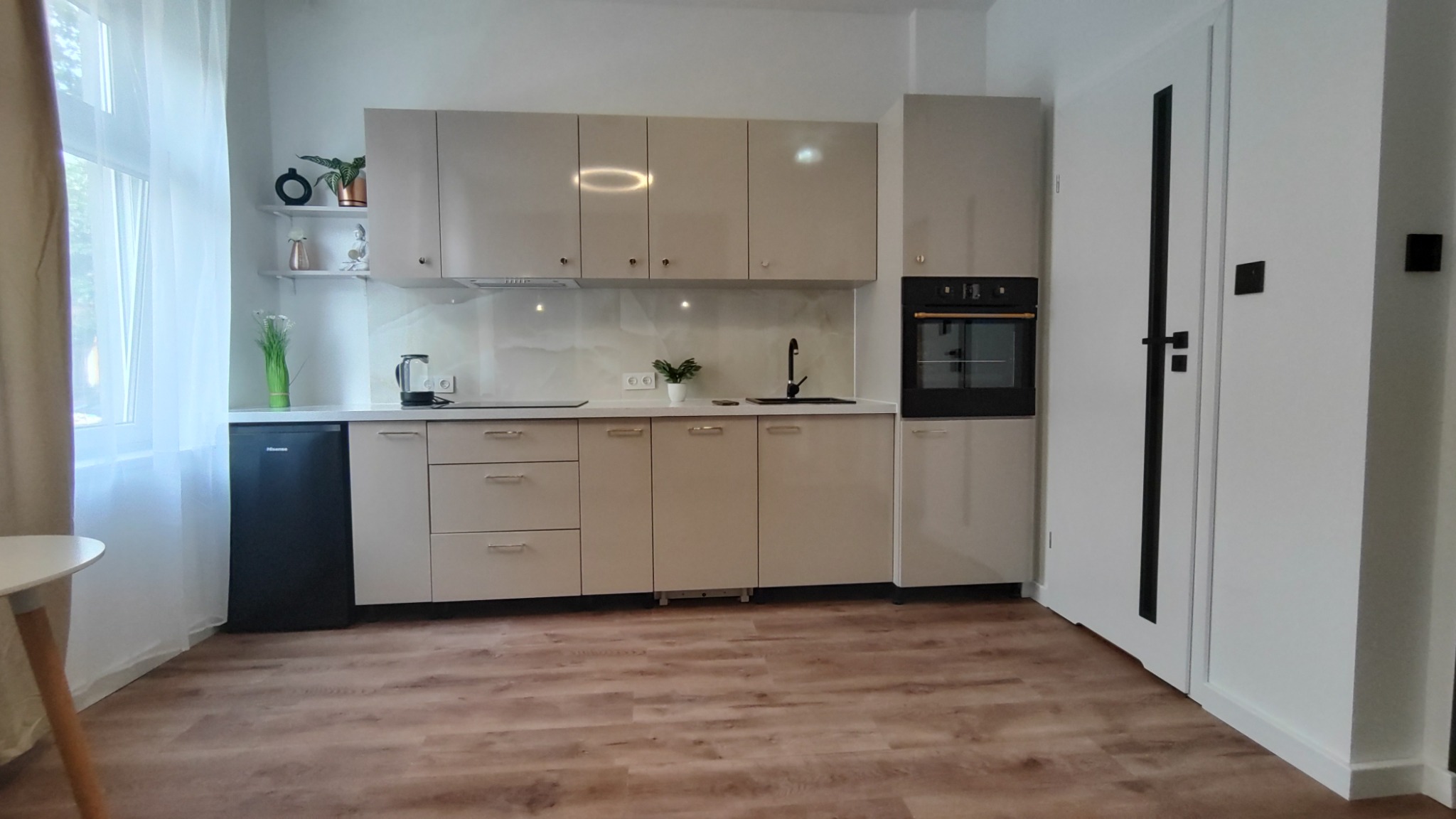 Jasna, nowoczesna kuchnia z beżowymi frontami, czarną armaturą i sprzętem AGD, uzupełniona o drewnopodobną podłogę i białe ściany z czarnymi akcentami.