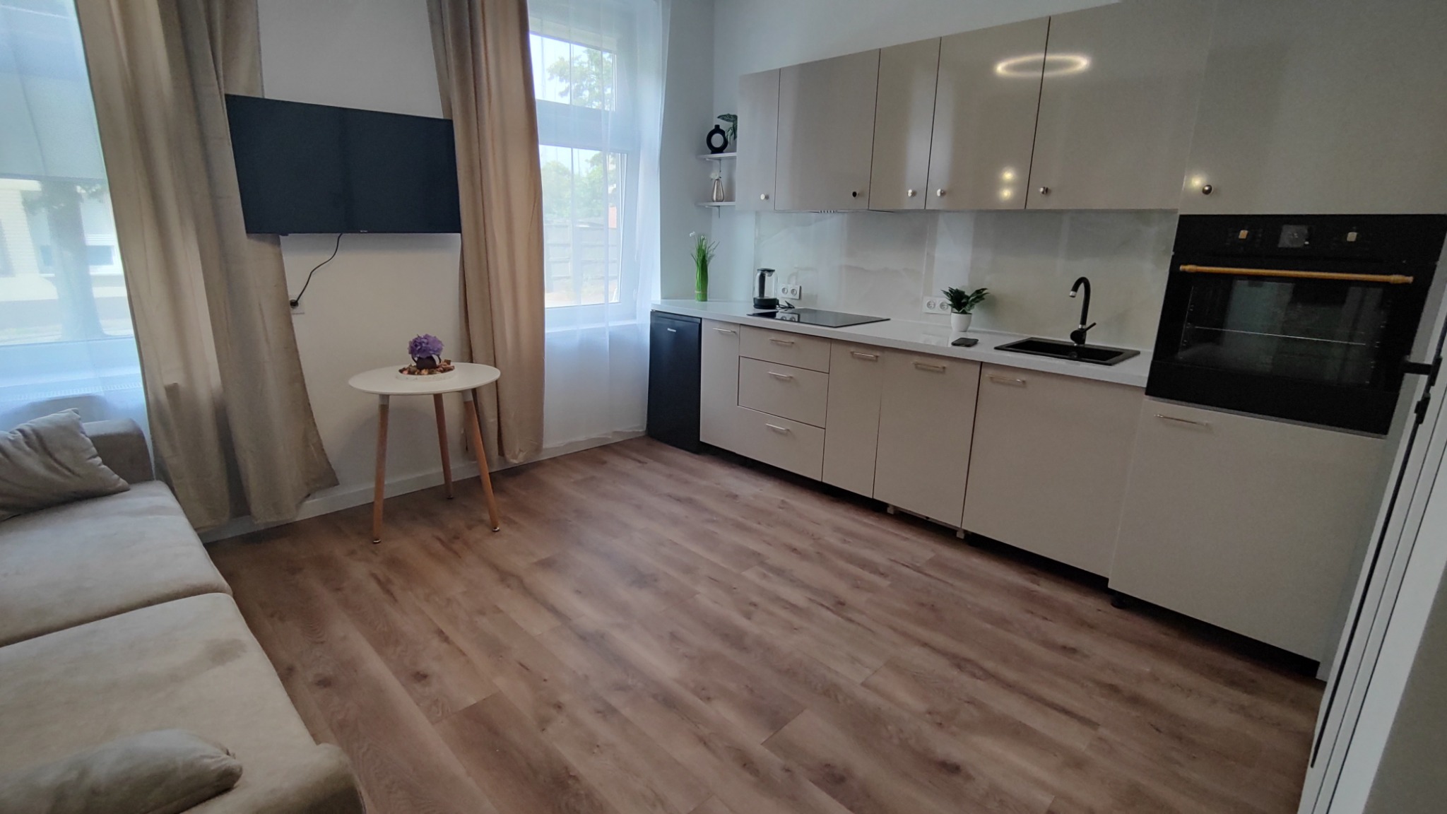 Jasna kuchnia z beżowymi frontami, czarnym piekarnikiem i płytą grzewczą, widok na drewnopodobną podłogę i fragment salonu z sofą i stolikiem.