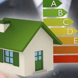 Model domu z zielonym dachem i oknami, obok diagram z klasami energetycznymi od A do G, trzymany przez osobę w garniturze.