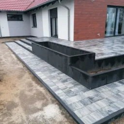 Nowo ułożona, wzorzysta kostka brukowa w odcieniach szarości na tarasie i ścieżce przy domu z elementami małej architektury, widoczna betoniarka i przygotowany teren.