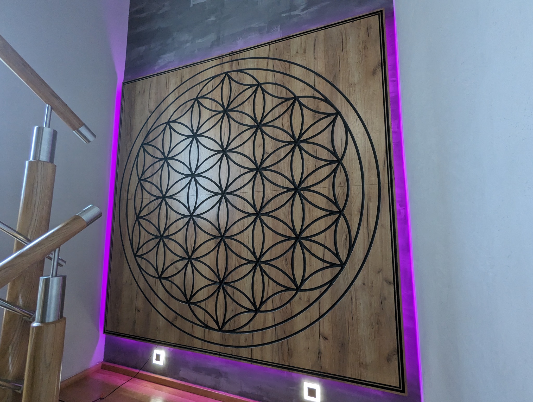 Drewniana ściana z wyciętym wzorem Kwiat Życia, podświetlona fioletowym światłem LED, widoczny fragment schodów z drewnianą balustradą.