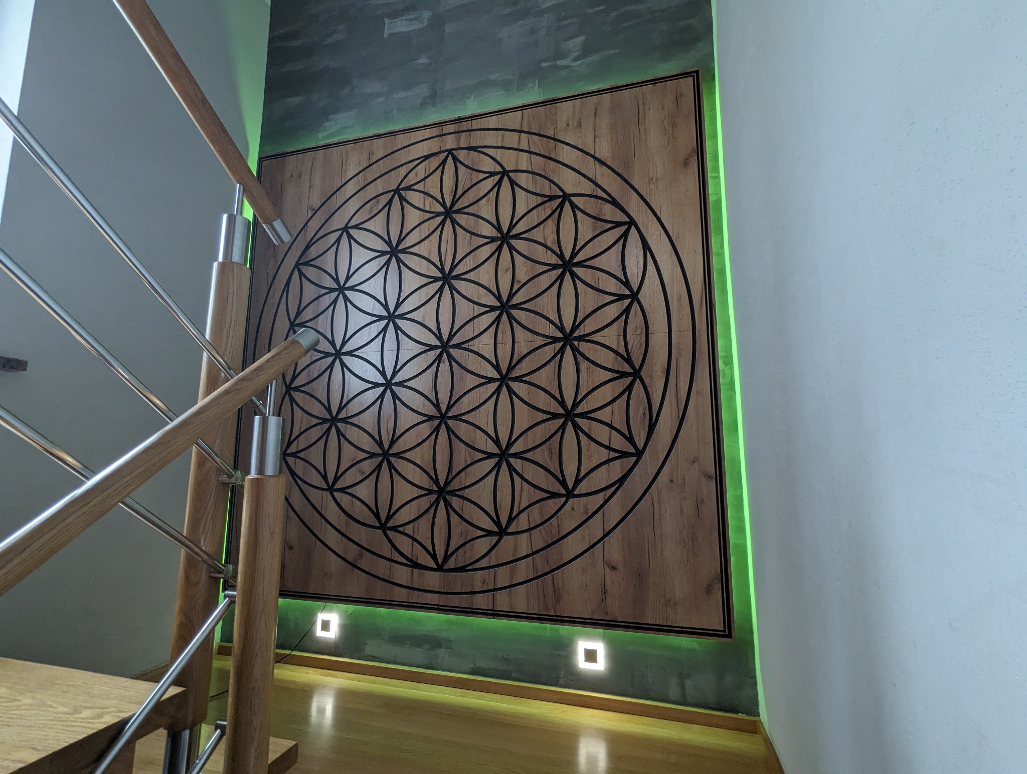 Drewniana ściana z geometrycznym wzorem kwiatu życia, podświetlona zielonym LED, widoczny fragment schodów z drewnianymi poręczami i metalowymi elementami.