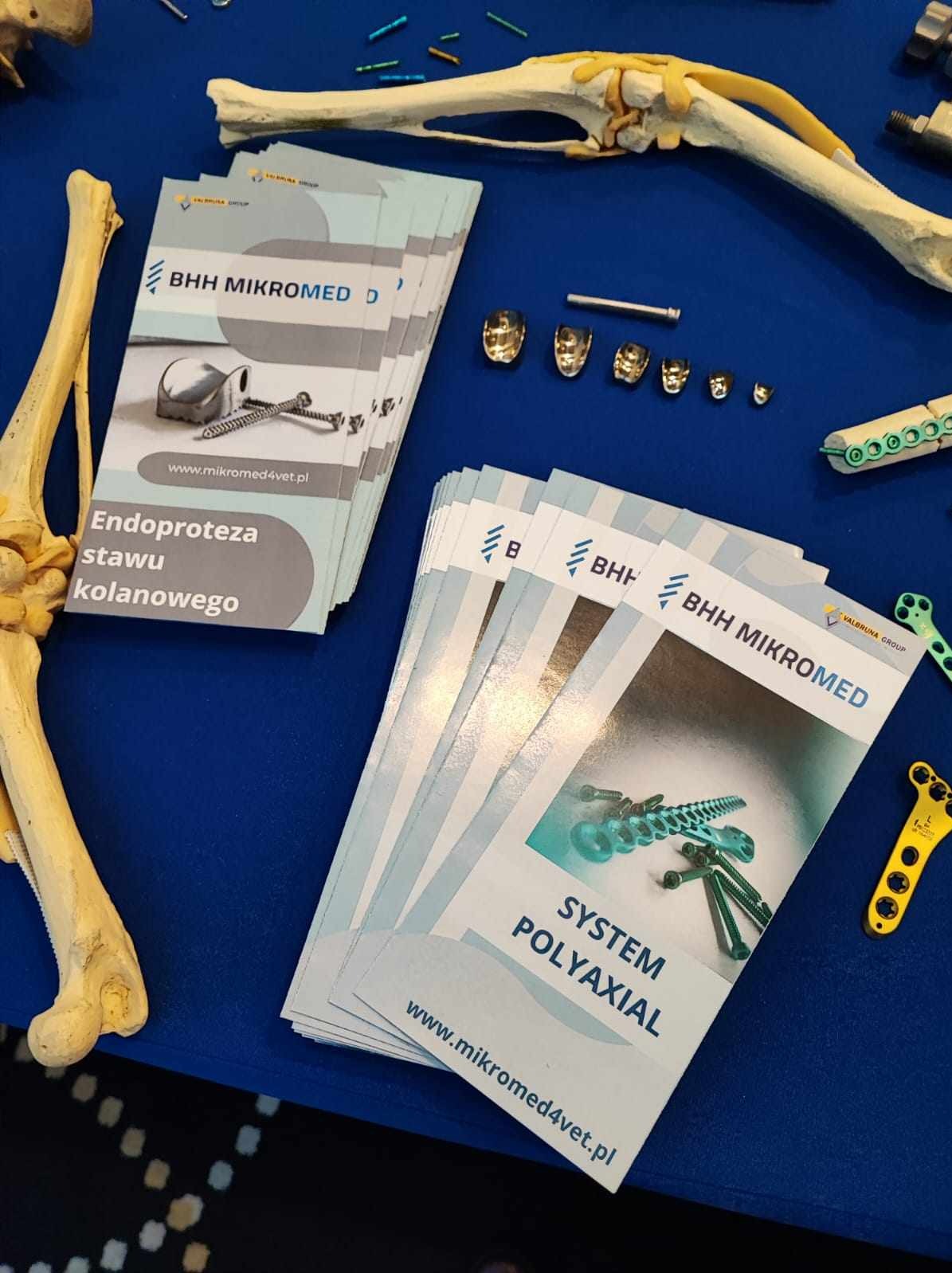Stoisko z broszurami i modelami kości oraz implantami firmy BHH Mikromed, prezentujące endoprotezy stawu kolanowego i systemy poliakselne na niebieskim tle.