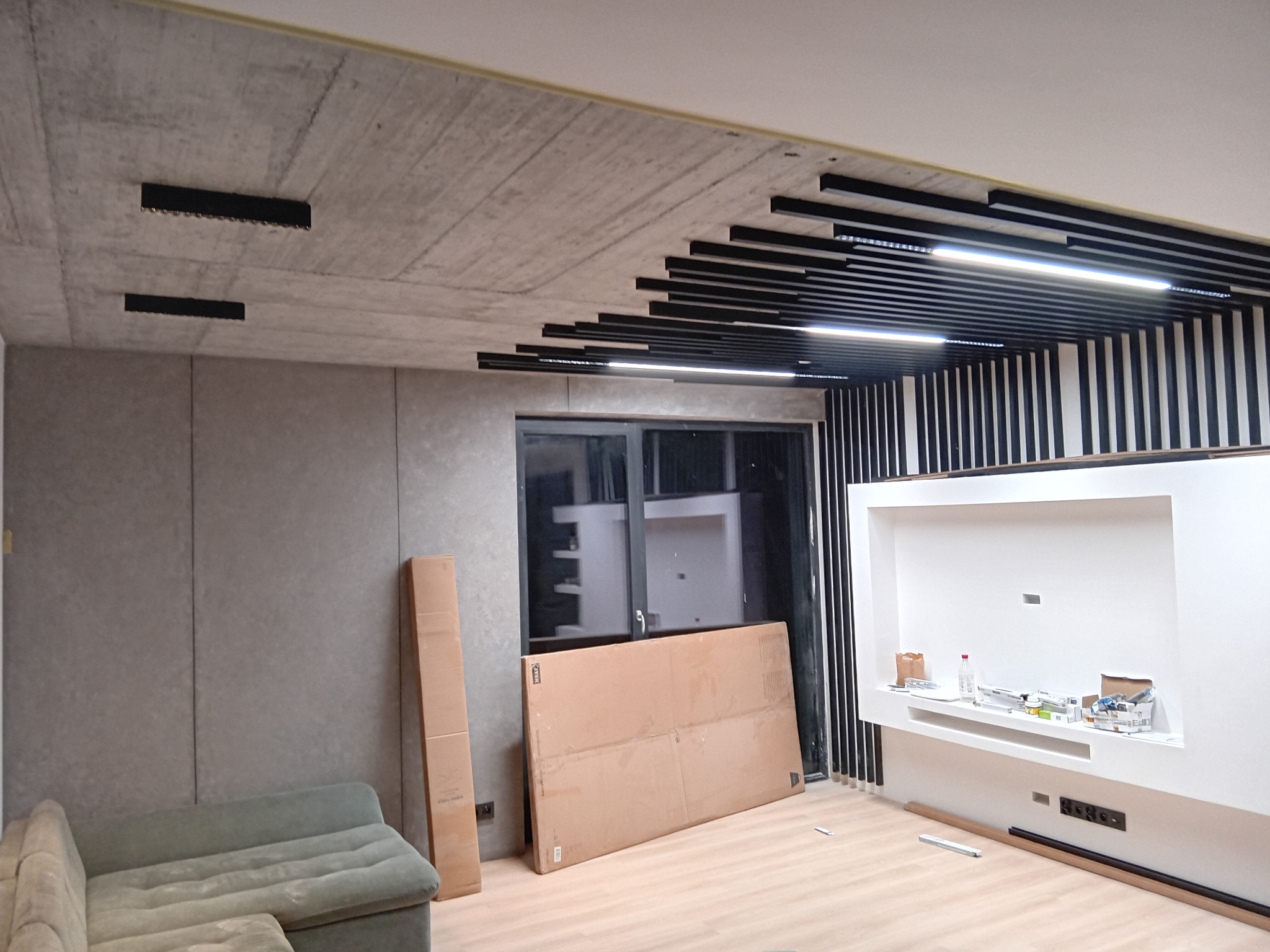 Wnętrze w trakcie wykończenia: sufit z lamelami i oświetleniem, ściana z efektem betonu, zabudowa pod telewizor, podłoga częściowo zakryta kartonami.
