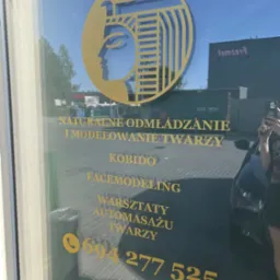 Złote logo na szklanych drzwiach salonu z profilem twarzy w stylu egipskim i napisem 'Naturalne odmładzanie i modelowanie twarzy, Kobido, Facemodeling, Warsztaty automasażu twarzy' oraz numerem...