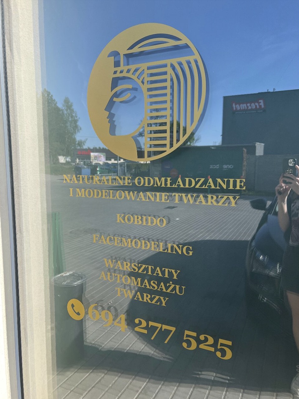 Złote logo na szklanych drzwiach salonu z profilem twarzy w stylu egipskim i napisem 'Naturalne odmładzanie i modelowanie twarzy, Kobido, Facemodeling, Warsztaty automasażu twarzy' oraz numerem...