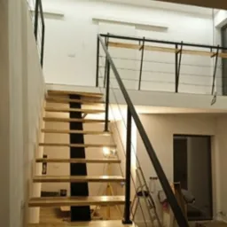 Nowoczesne schody drewniane z czarną metalową konstrukcją nośną i balustradą z linek, w jasnym wnętrzu domu z antresolą.