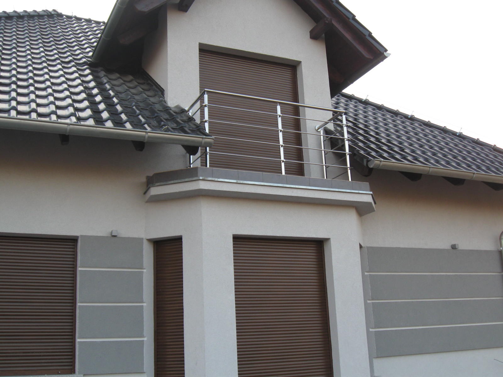 Balkon z nowoczesną, poziomą balustradą ze stali nierdzewnej, brązowe rolety zewnętrzne, elewacja w odcieniach szarości i bieli.