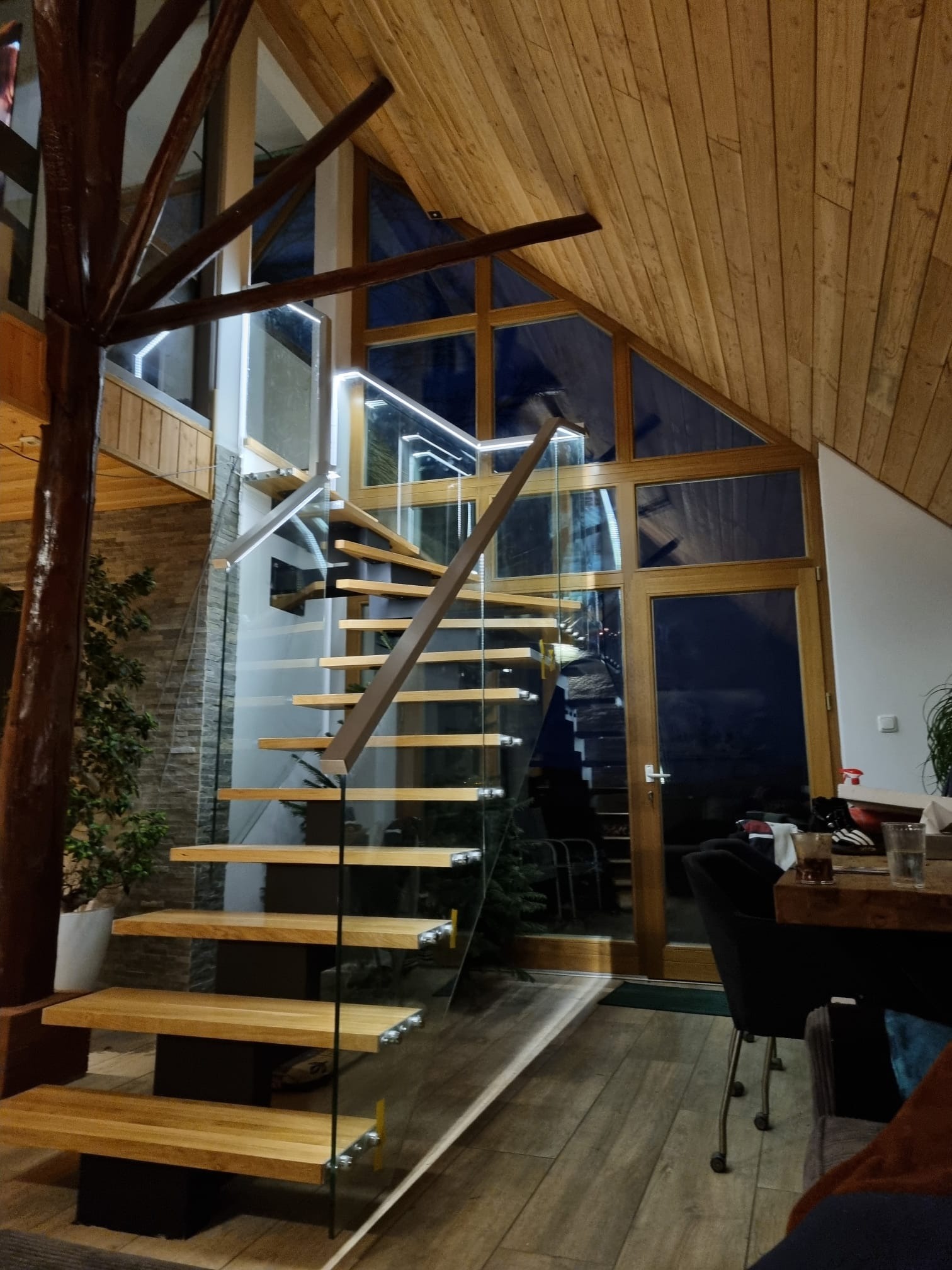 Nowoczesne schody z drewnianymi stopniami, szklanymi balustradami i oświetleniem LED w przestronnym wnętrzu z drewnianym sufitem i dużymi oknami.