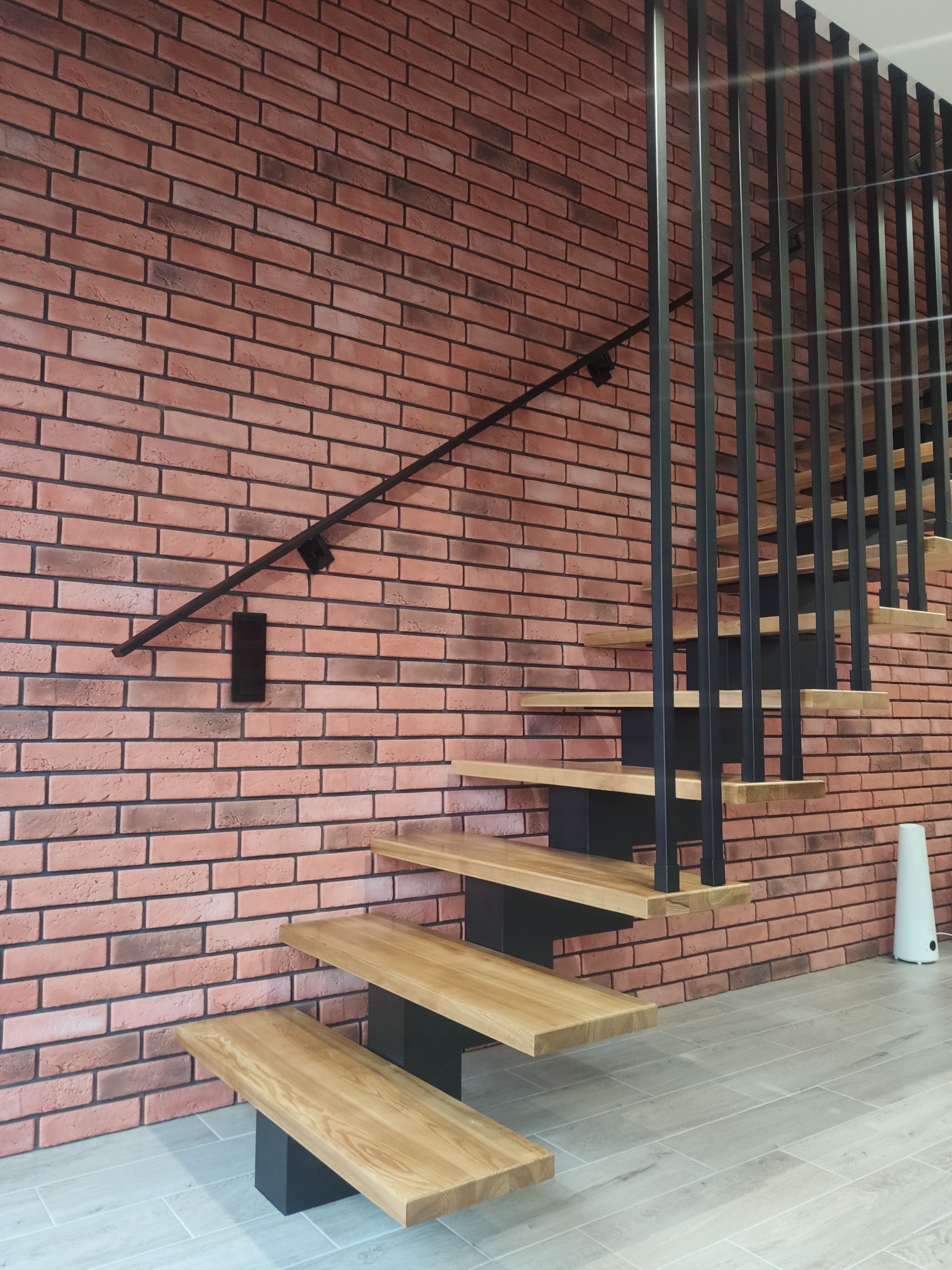 Nowoczesne schody z drewnianymi stopniami i czarną metalową balustradą na tle ściany z cegły.