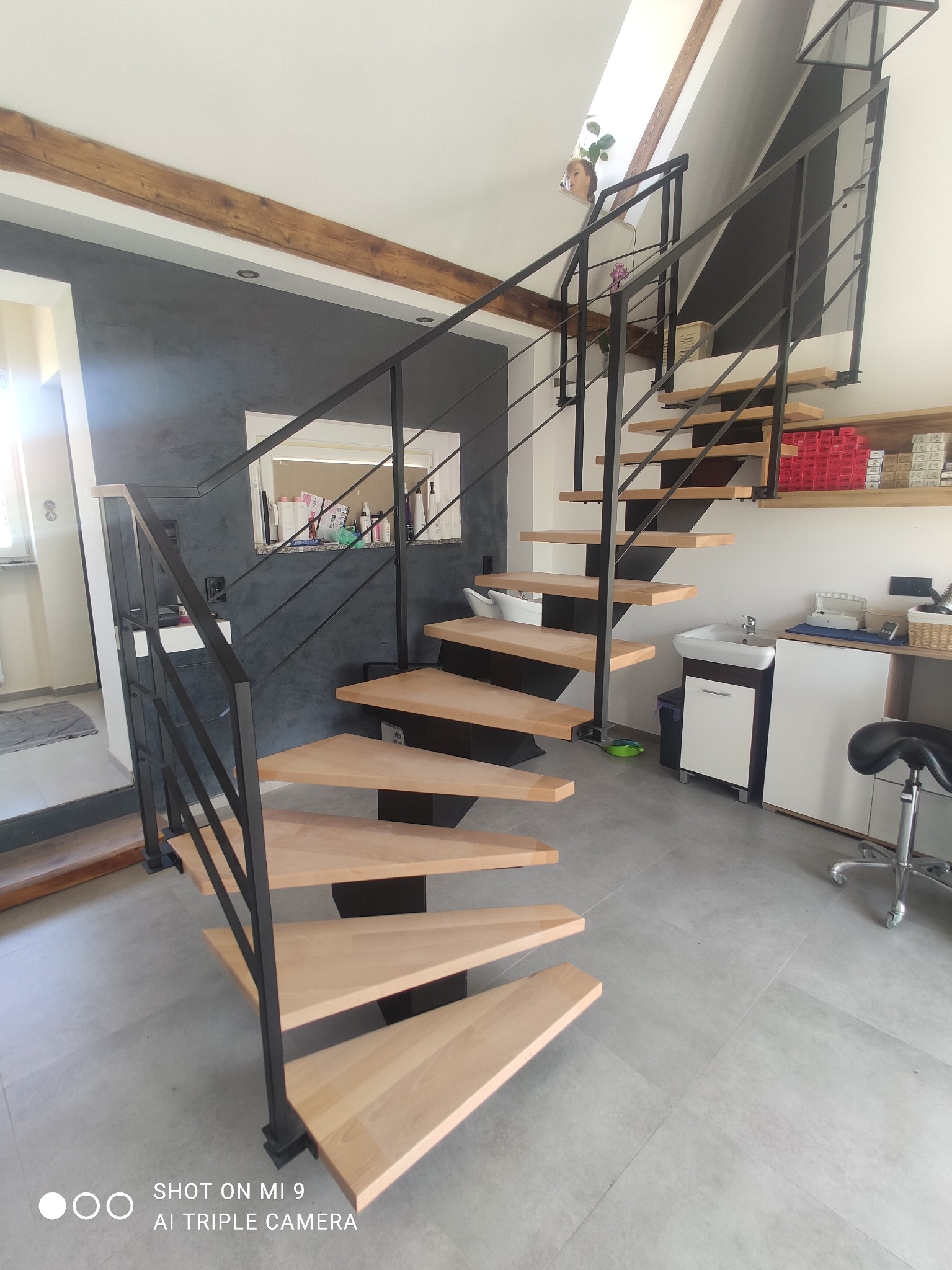 Spiralne schody z drewnianymi stopniami i czarną metalową balustradą w jasnym wnętrzu z elementami rustykalnymi.