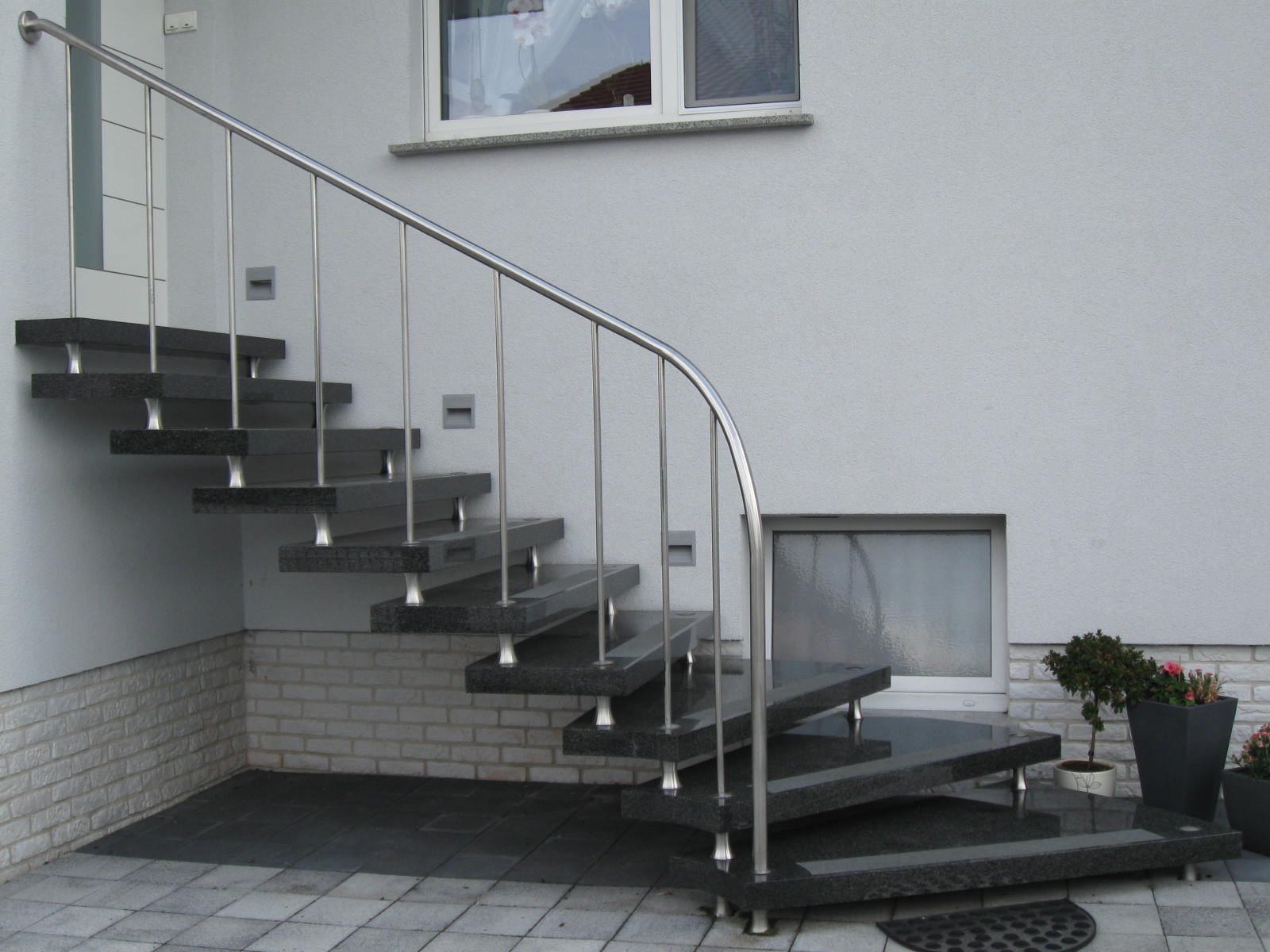 Zewnętrzne schody z czarnego kamienia z balustradą ze stali nierdzewnej, wsparte na metalowych nóżkach, prowadzące do drzwi wejściowych.