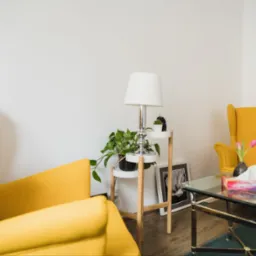 Ustawienie pokoju psychoterapeuty: dwa żółte fotele uszakowe, szklany stolik kawowy z kwiatami i pudełkiem chusteczek, lampa na stoliku pomocniczym z rośliną doniczkową i czarno-białe zdjęcie w ramce.