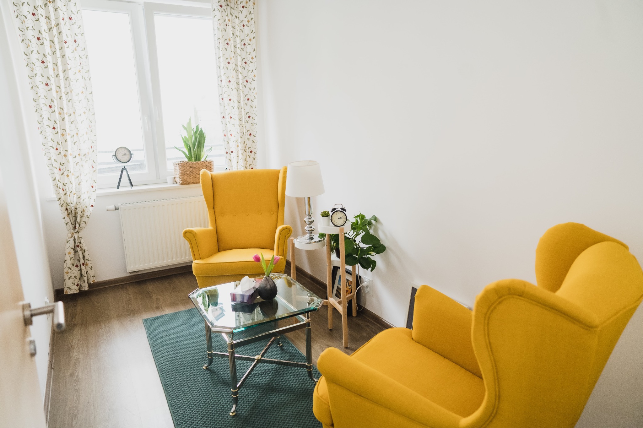 Jasne wnętrze gabinetu z dwoma żółtymi fotelami typu uszak, stolikiem kawowym ze szklanym blatem, lampą stołową, zegarem i roślinami doniczkowymi, tworzące przytulną i profesjonalną atmosferę.